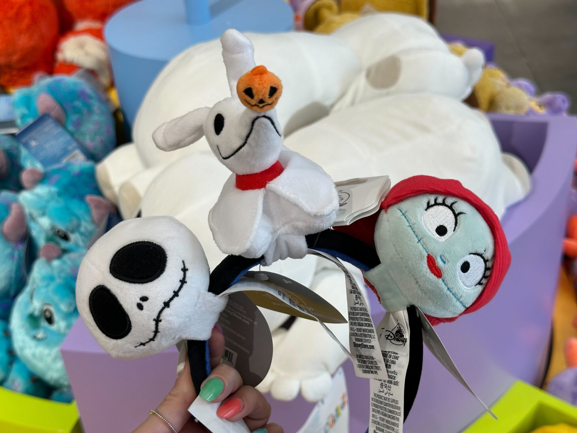 A person holds a headband featuring plush characters from "The Nightmare Before Christmas": Jack Skellington, Zero the ghost dog, and Sally. Stuffed toys are displayed in the background.