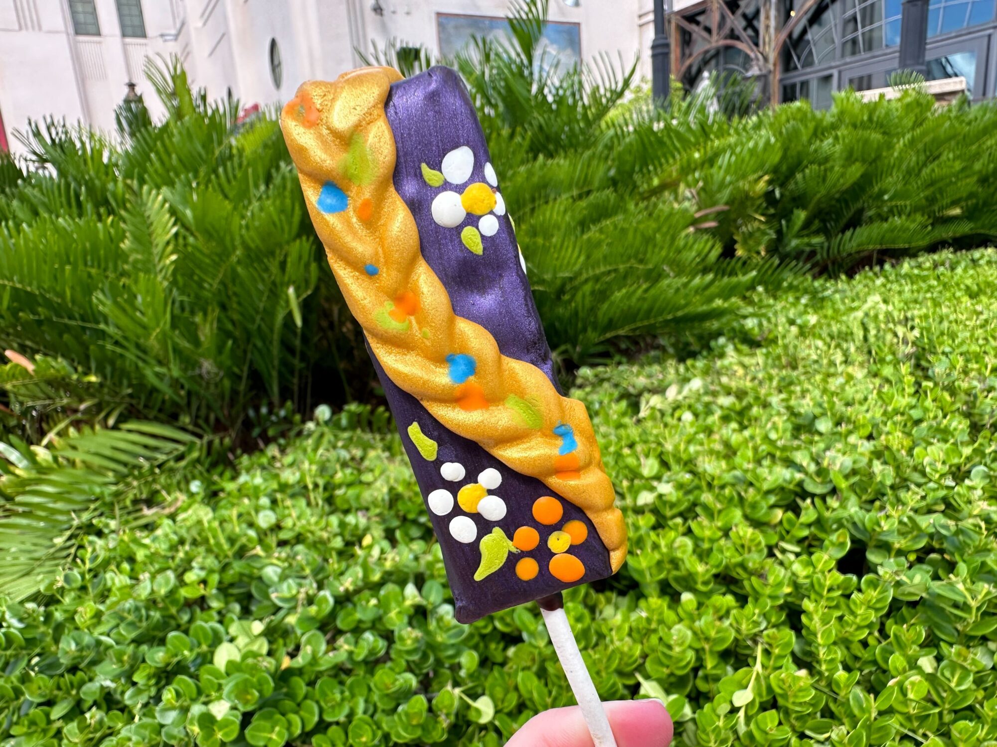 A decorative rectangular popsicle with colorful designs is held up against a background of lush green foliage and buildings.