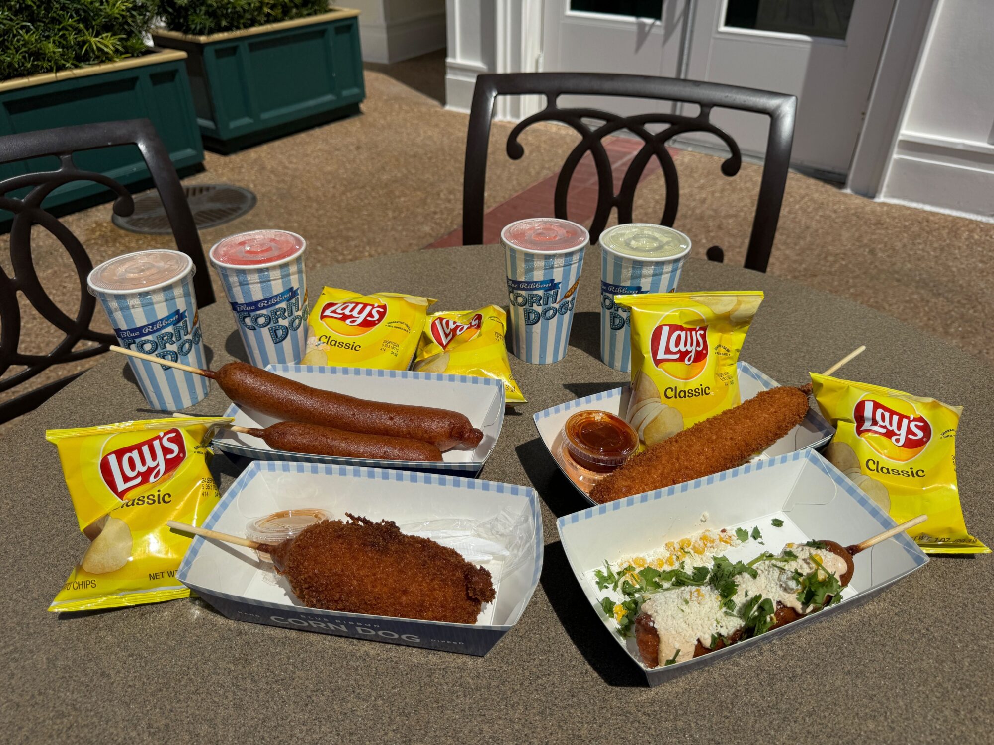 A table with four servings of corndogs and Lay's Classic potato chips, four drinks in "Corn Dog" cups, and dipping sauces, set in an outdoor seating area.