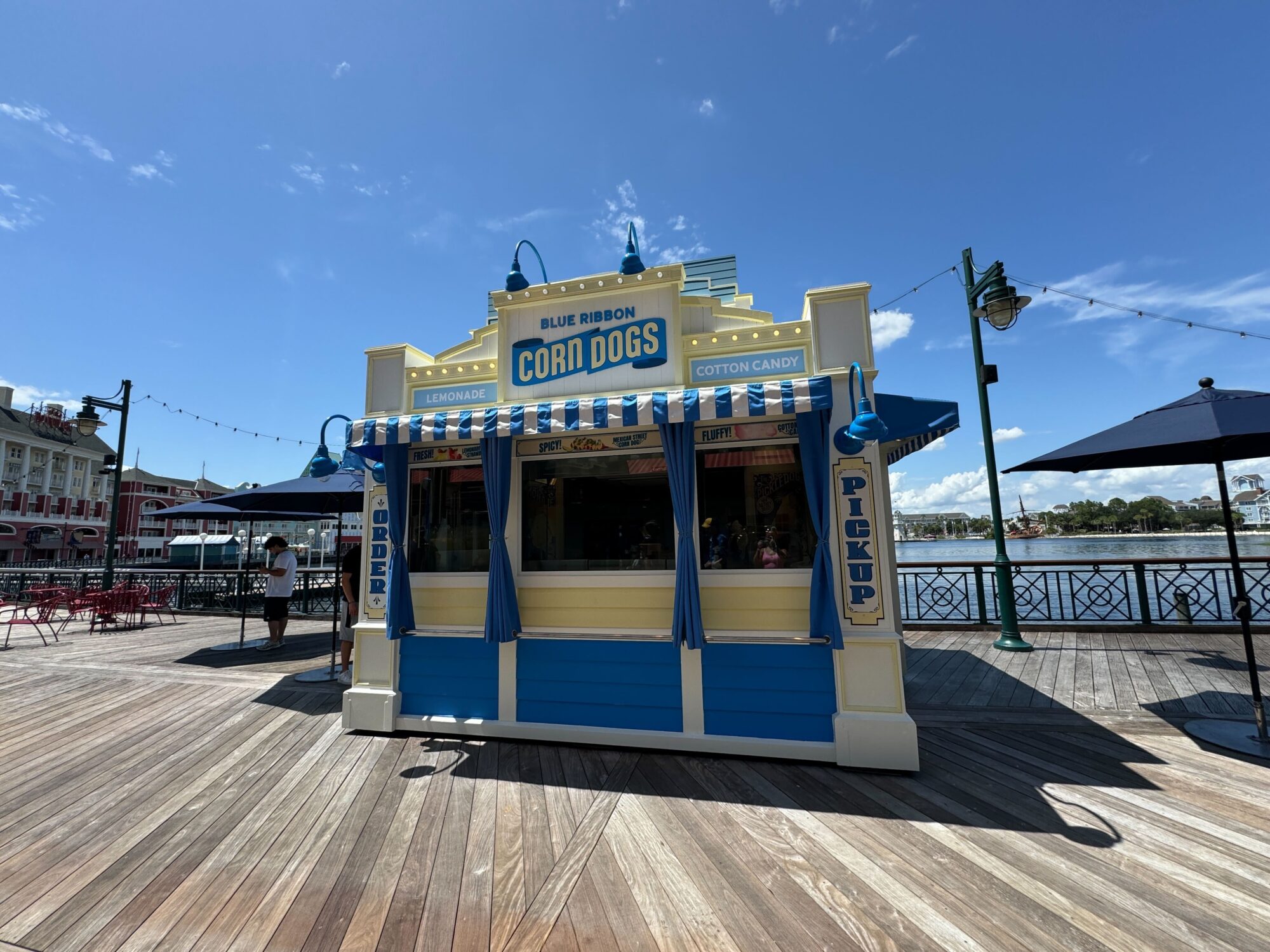 A blue and yellow food stand on a boardwalk selling corn dogs and cotton candy. The stand has "Blue Ribbon Corn Dogs" signage and is next to a body of water with outdoor seating nearby.
