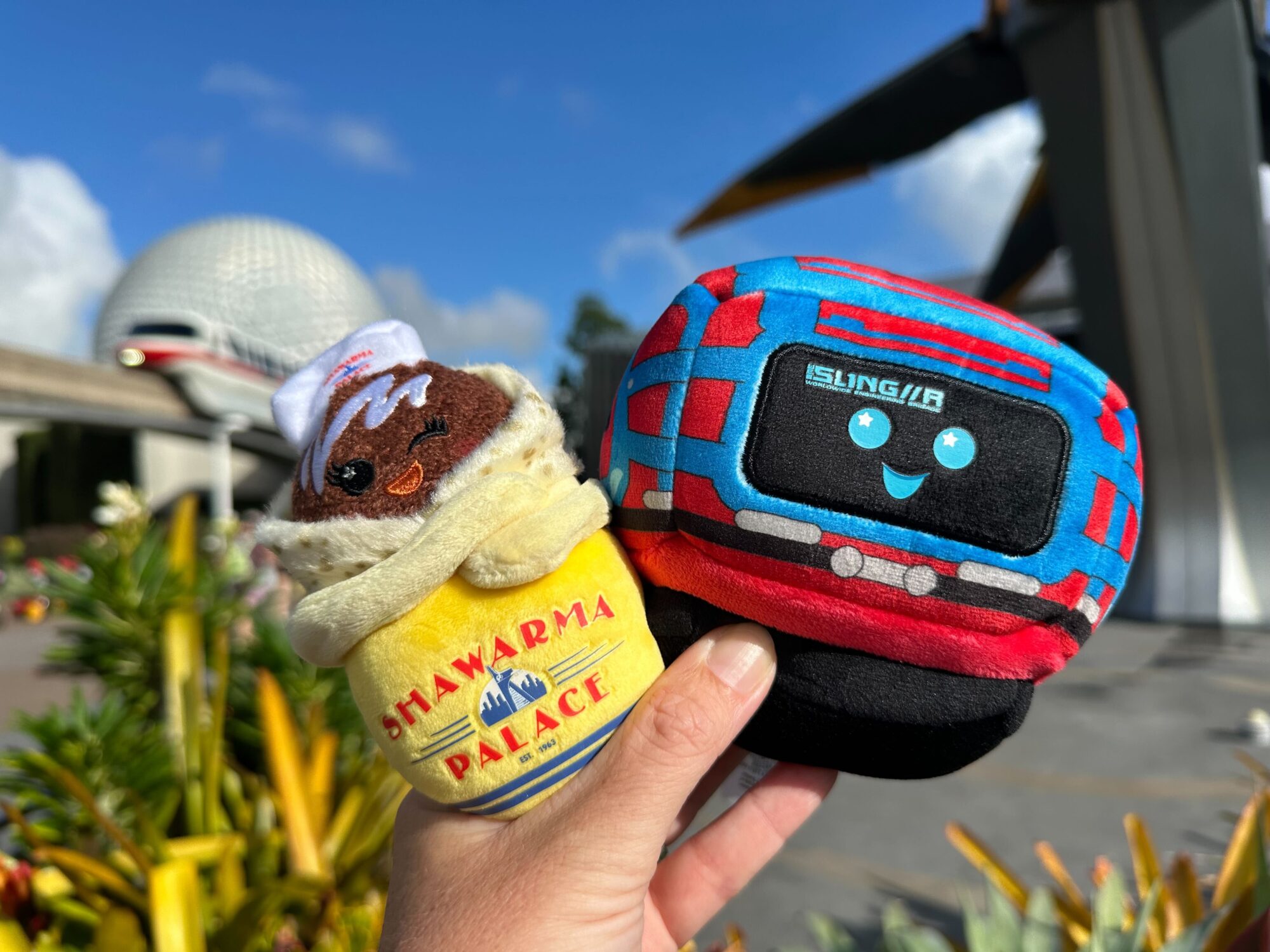 A hand holds two plush toys in front of Spaceship Earth at Epcot. One toy is a rolled shawarma with a "Shawarma Palace" wrap, and the other is a smiling toy bus with "SLING//R" on it.