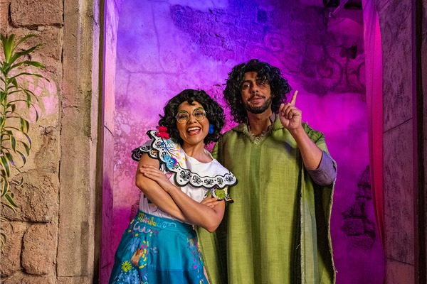 Two people dressed as Mirabel and Bruno from the movie "Encanto" pose in front of a stone wall with purple lighting at Mickey's Not-So-Scary Halloween Party. One person smiles with arms crossed, while the other raises a finger and looks ahead.