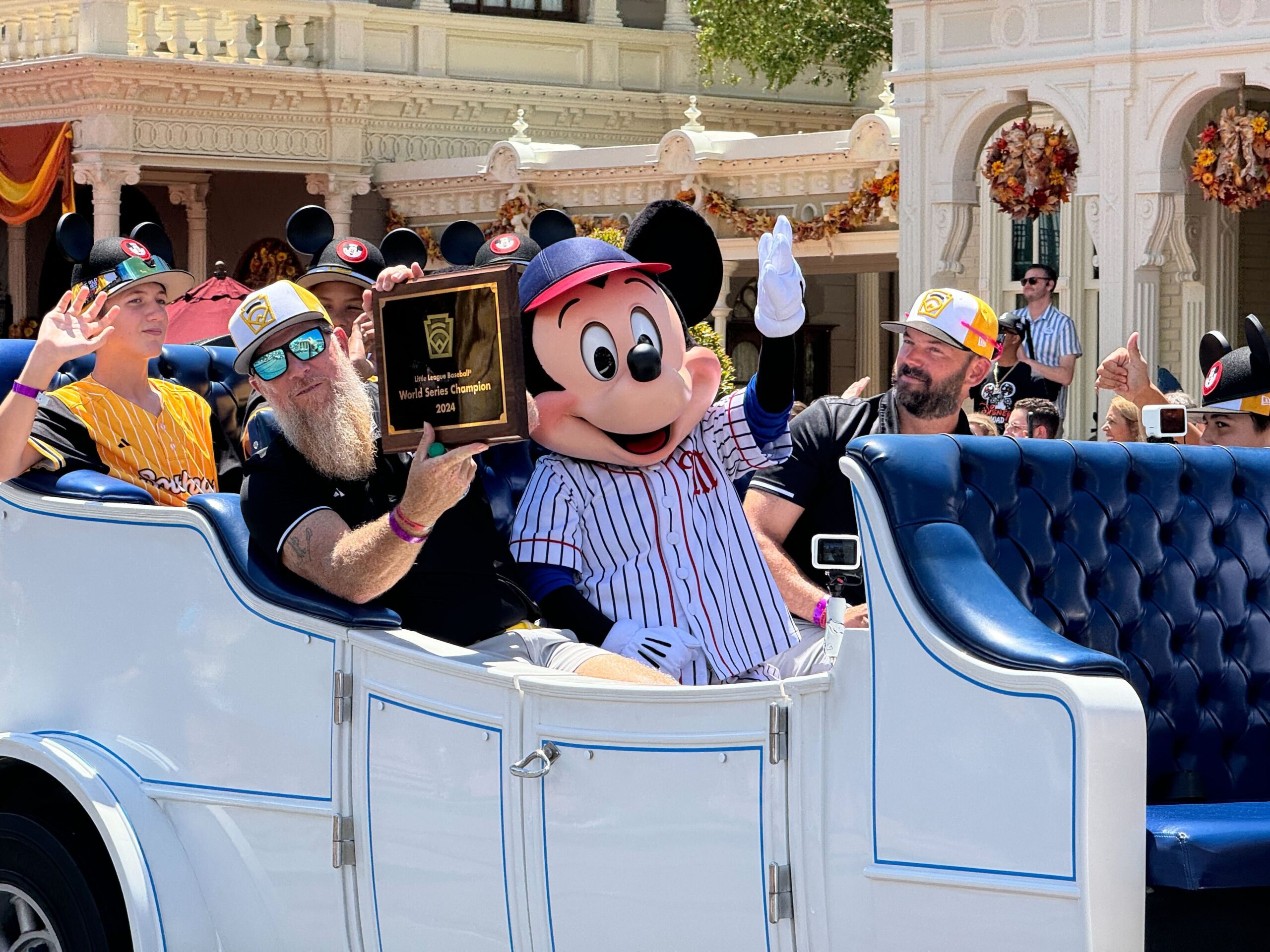 A group of champions in a white carriage, one holding an award plaque, accompanied by someone in a Mickey Mouse costume wearing a baseball uniform, wave at the camera in a festive Magic Kingdom setting.