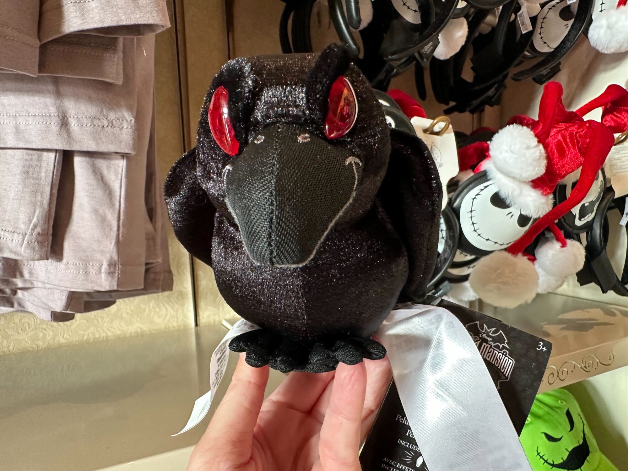 Close-up of a black plush toy with red eyes being held in front of a display featuring stuffed figures with white faces and Santa hats, reminiscent of the Haunted Mansion's eerie charm.