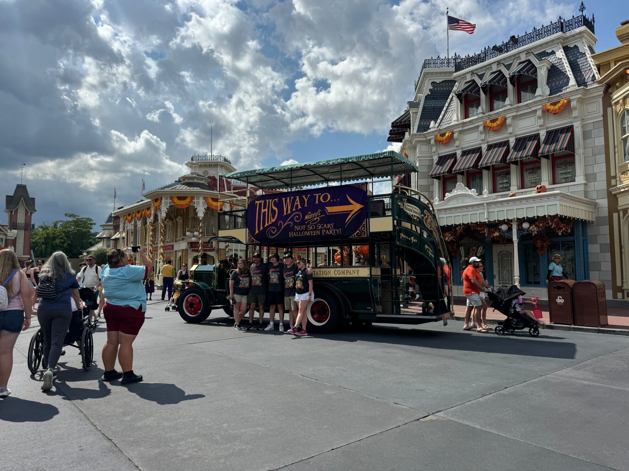 A group of people stands next to a vintage bus with a sign that reads "This way to... Mickey's Not-So-Scary Halloween Party 2024" on a sunny day in Magic Kingdom, with ornate buildings in the background.