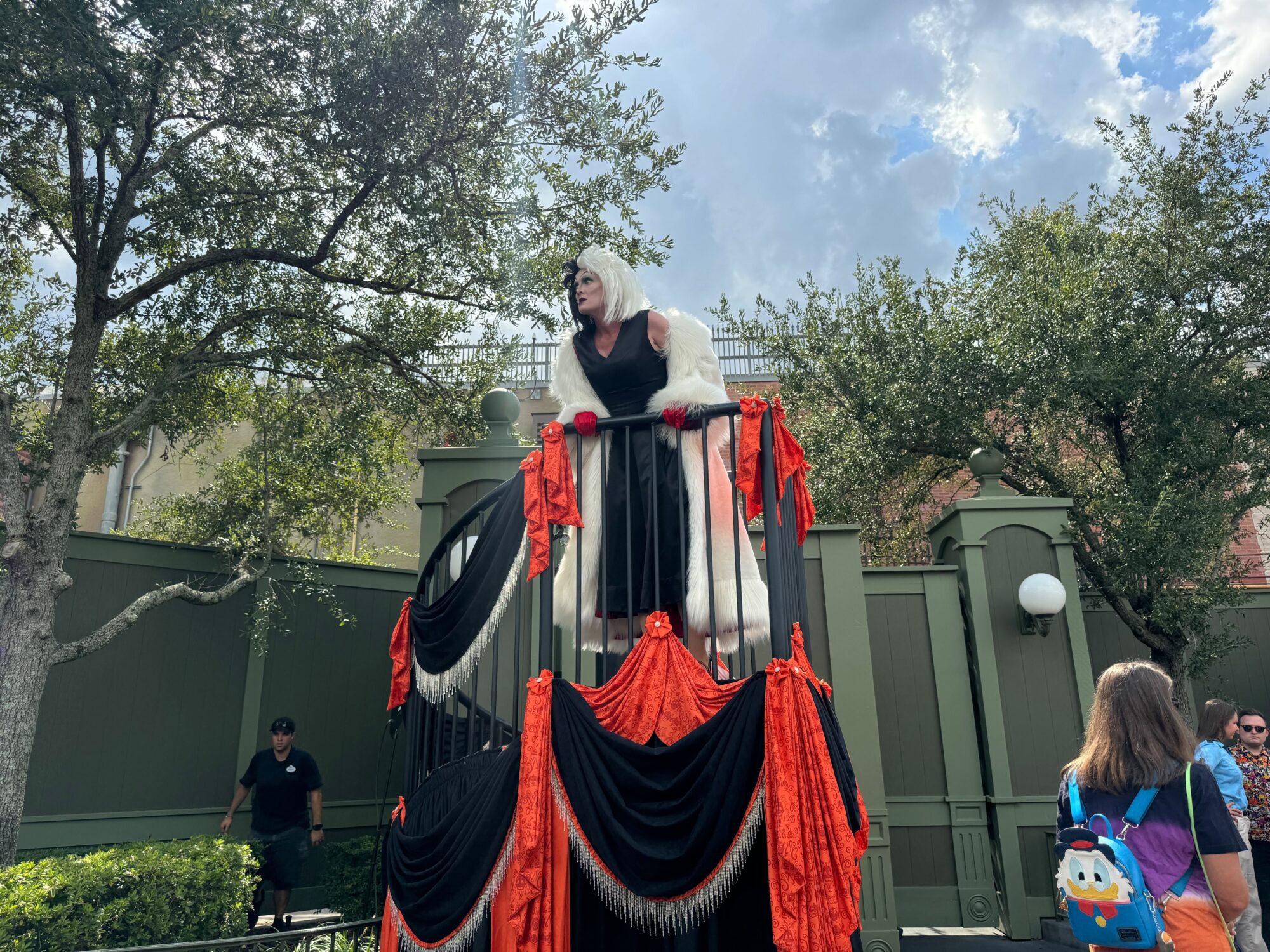 A person dressed in a black and white costume stands atop a black and red decorated platform, surrounded by trees. An individual with a blue backpack is observing from below, as if following a full guide through the not-so-scary Halloween party.