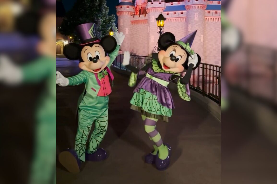 Two costumed characters resembling mice dressed in green and purple Halloween outfits pose in front of a pink castle at night.