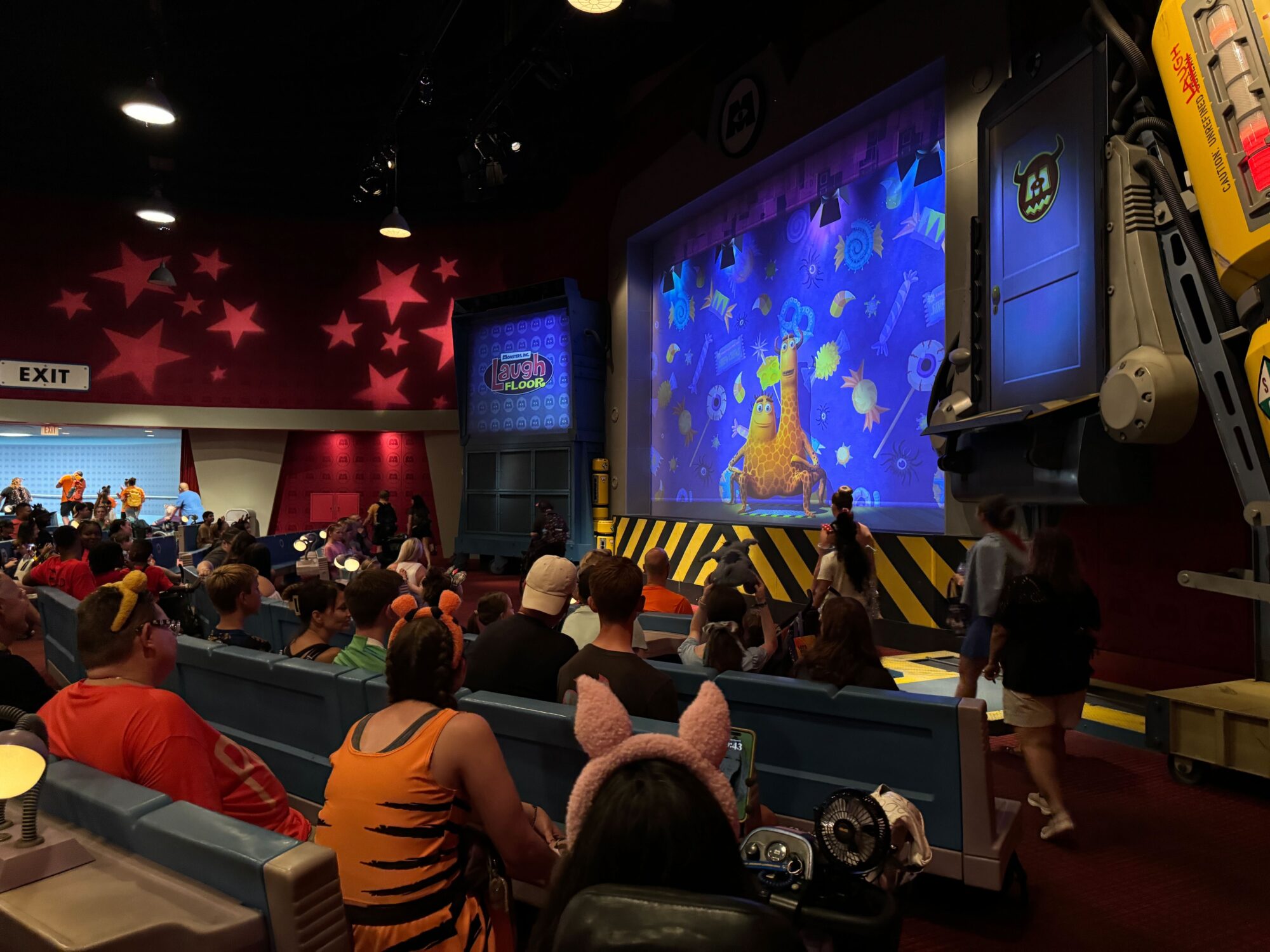 A group of people seated in a theater-style room at Mickey’s Not-So-Scary Halloween Party in Magic Kingdom watches an animated character on a large screen. The room has themed decorations and colorful lighting, adding to the festive 2024 atmosphere.