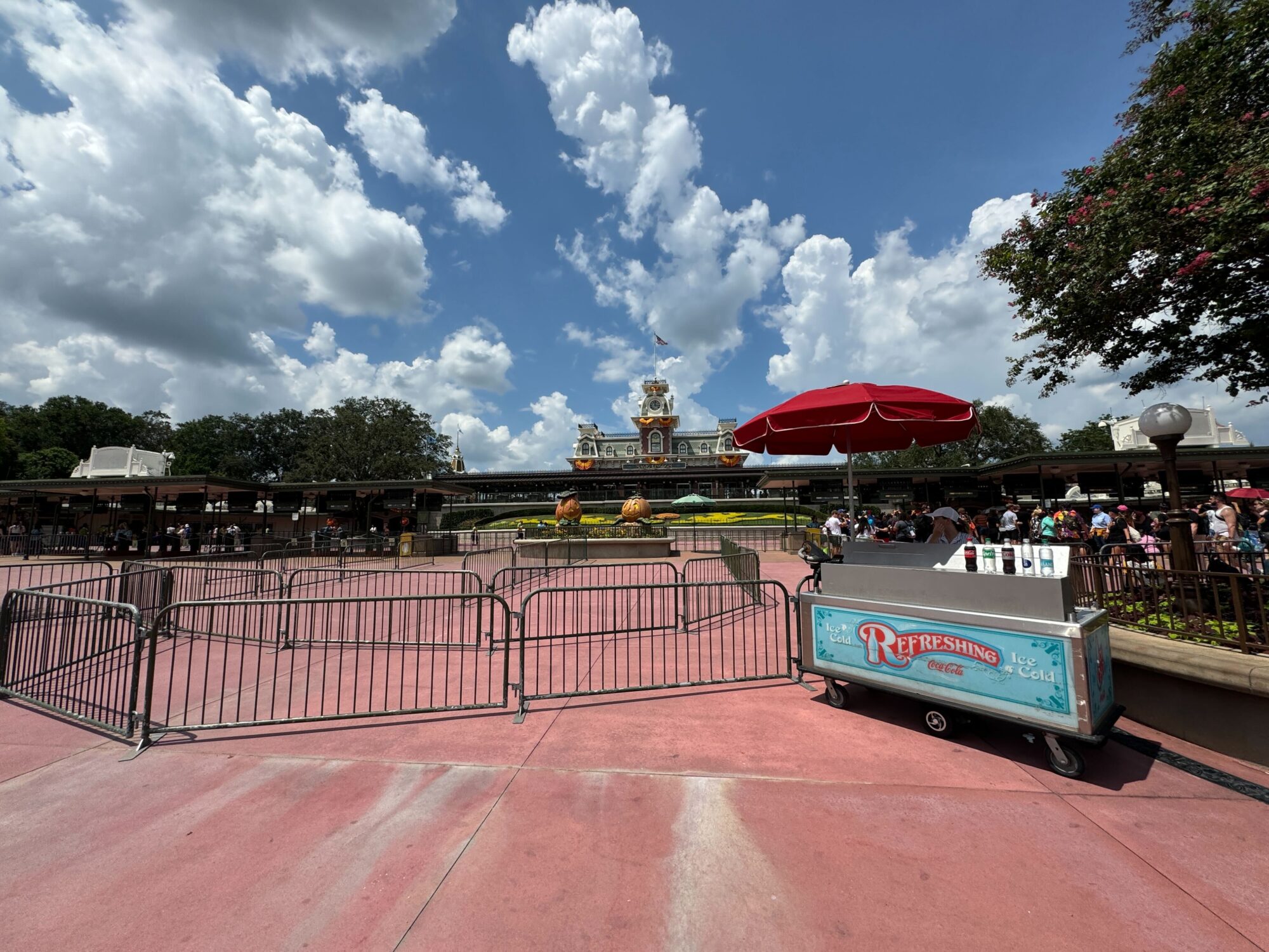 An entrance area with a red umbrella and metal barricades, and a cart labeled "Refreshing Ice Cold Drinks." In the background is a building with a clock tower and decorative landscaping.