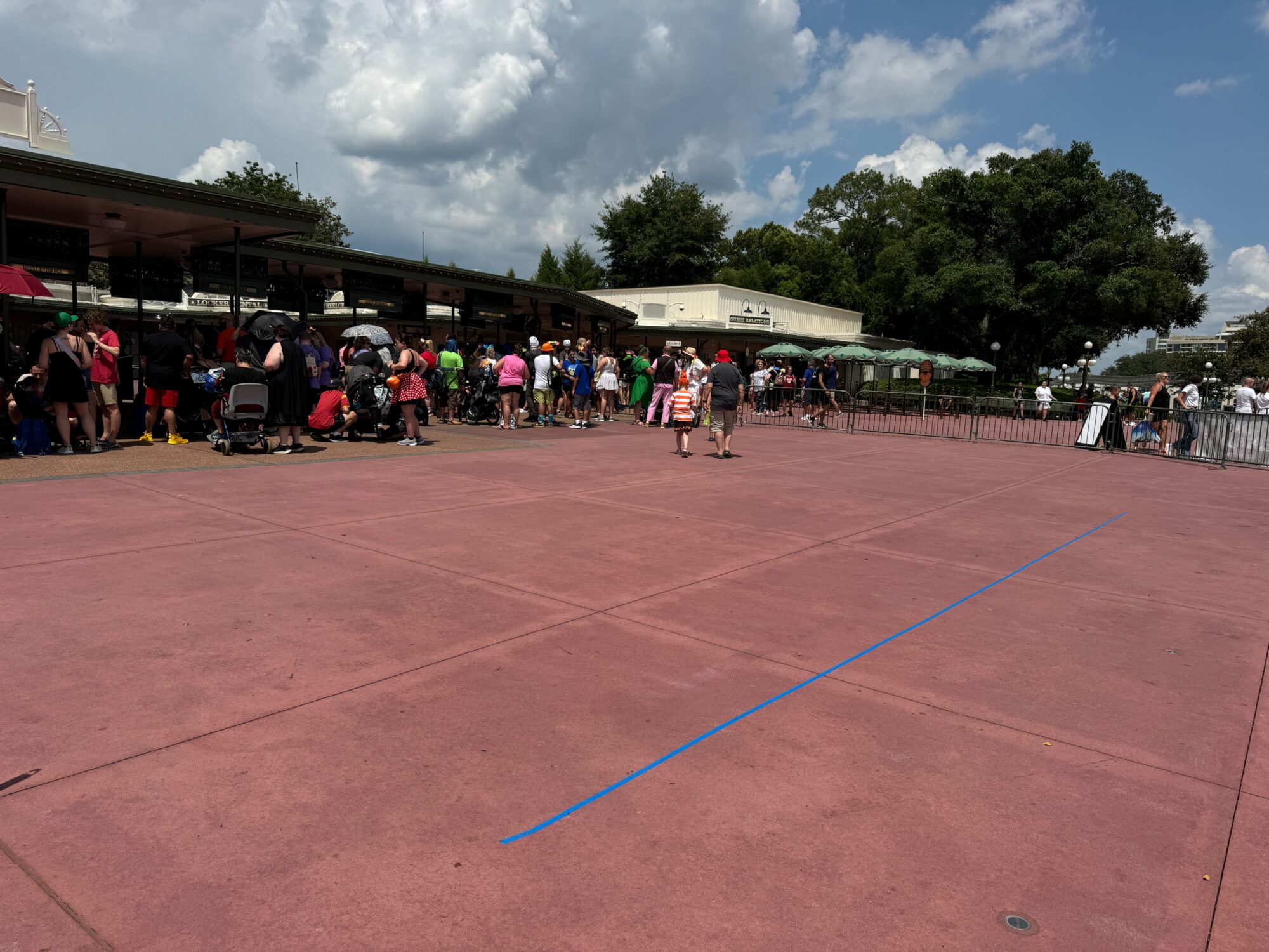 A crowd of people stands in line outside a building under a cloudy sky. A tree is in the background, and blue tape marks the ground in the foreground.