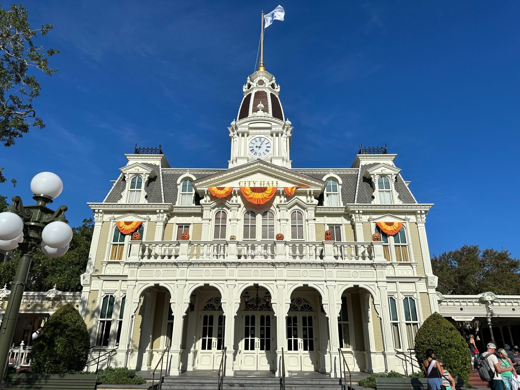 A large, ornate building with a clock tower labeled "City Hall" decorated with yellow and orange bunting against a clear blue sky. People are seen at the entrance and walking nearby.