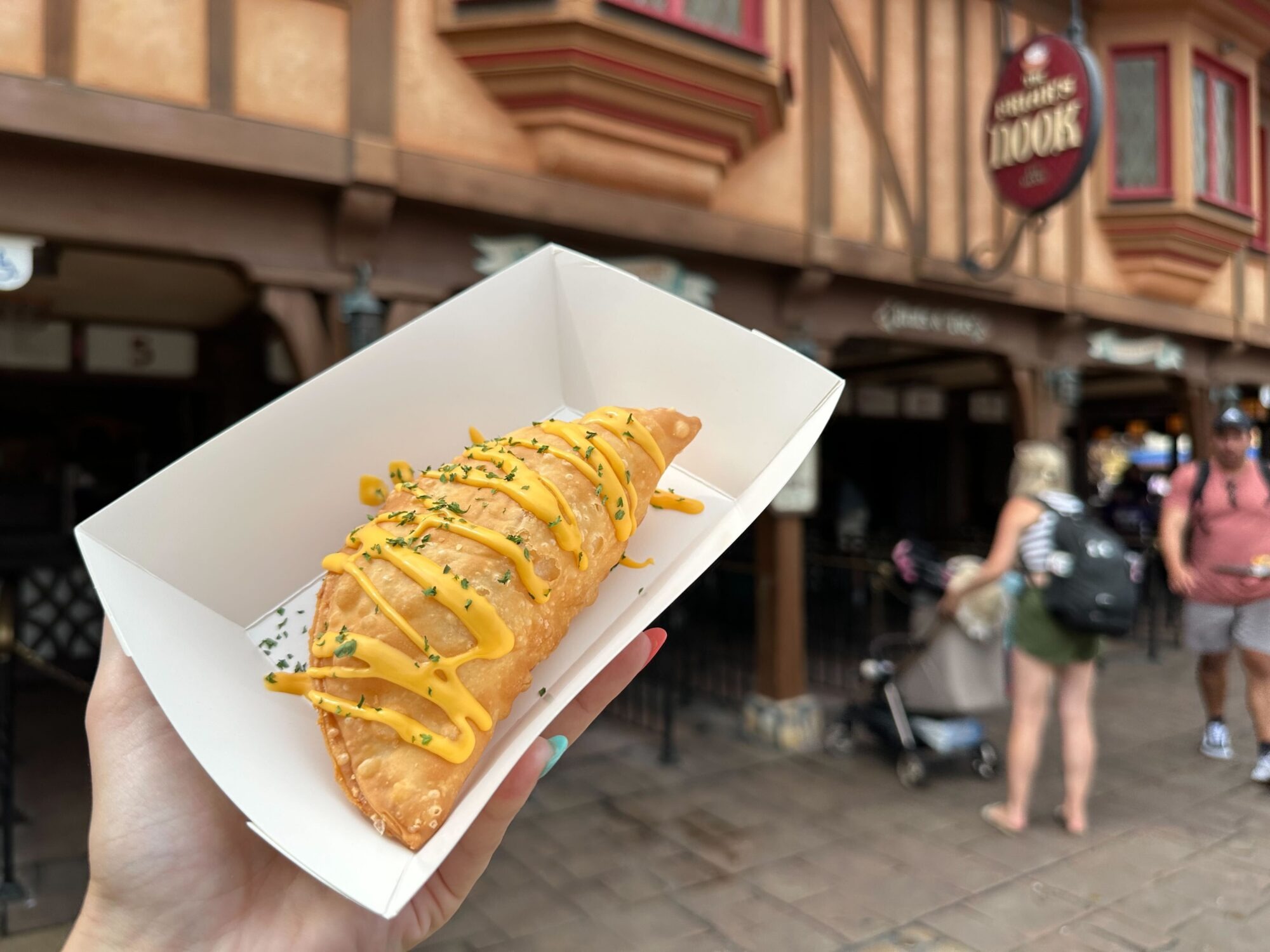 A person holds a fried hand pie topped with a yellow sauce and green herbs in front of a Tudor-style building with a "Croque Glace" sign. Two people and a stroller are visible in the background, making this an intriguing scene for any food review enthusiast.