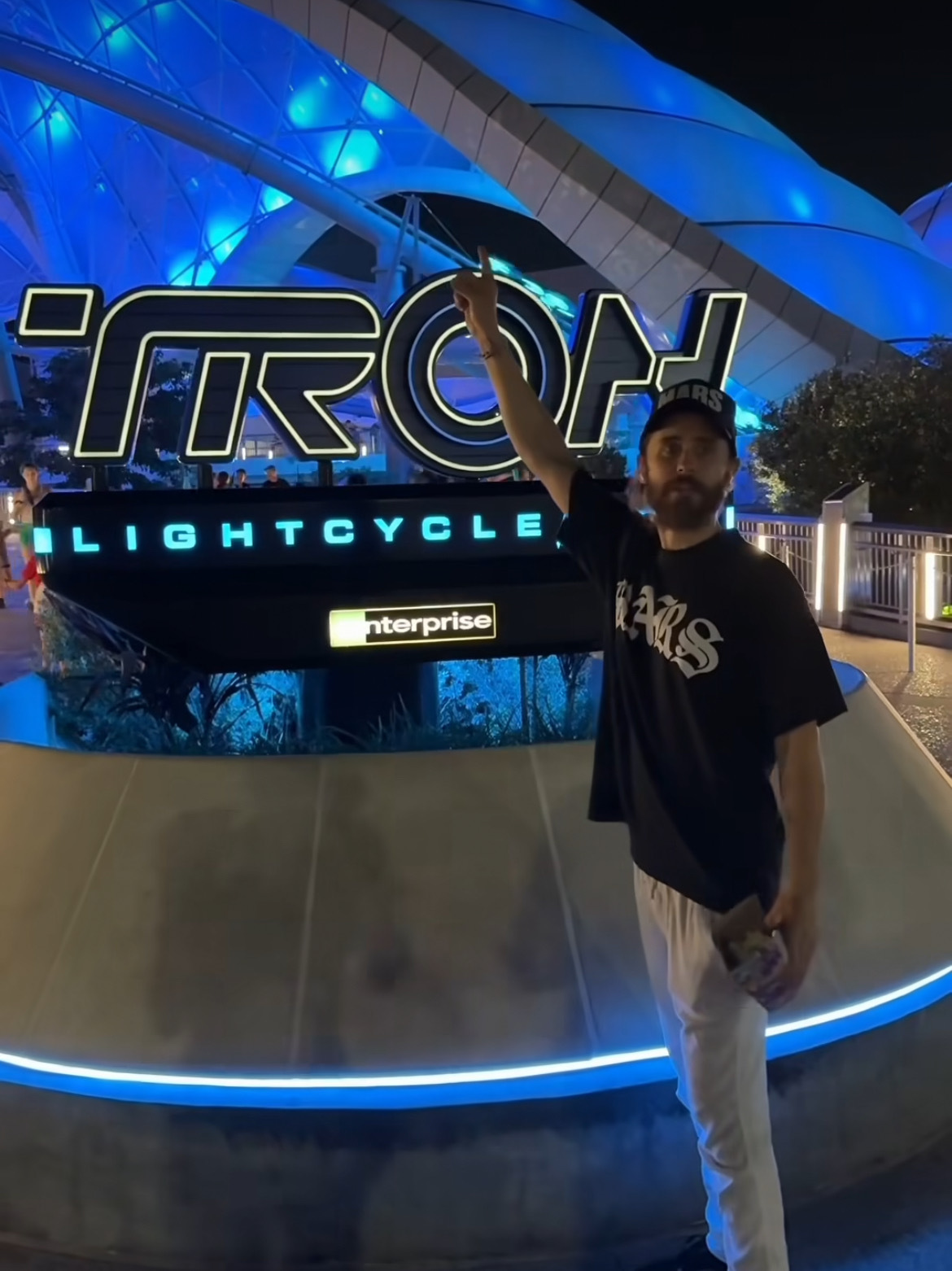 A man in a black shirt and white pants points up at a sign for "TRON Lightcycle Power Run" in front of a futuristic, illuminated blue structure at night. You could almost imagine Jared Leto stepping into this scene from one of his sci-fi films.
