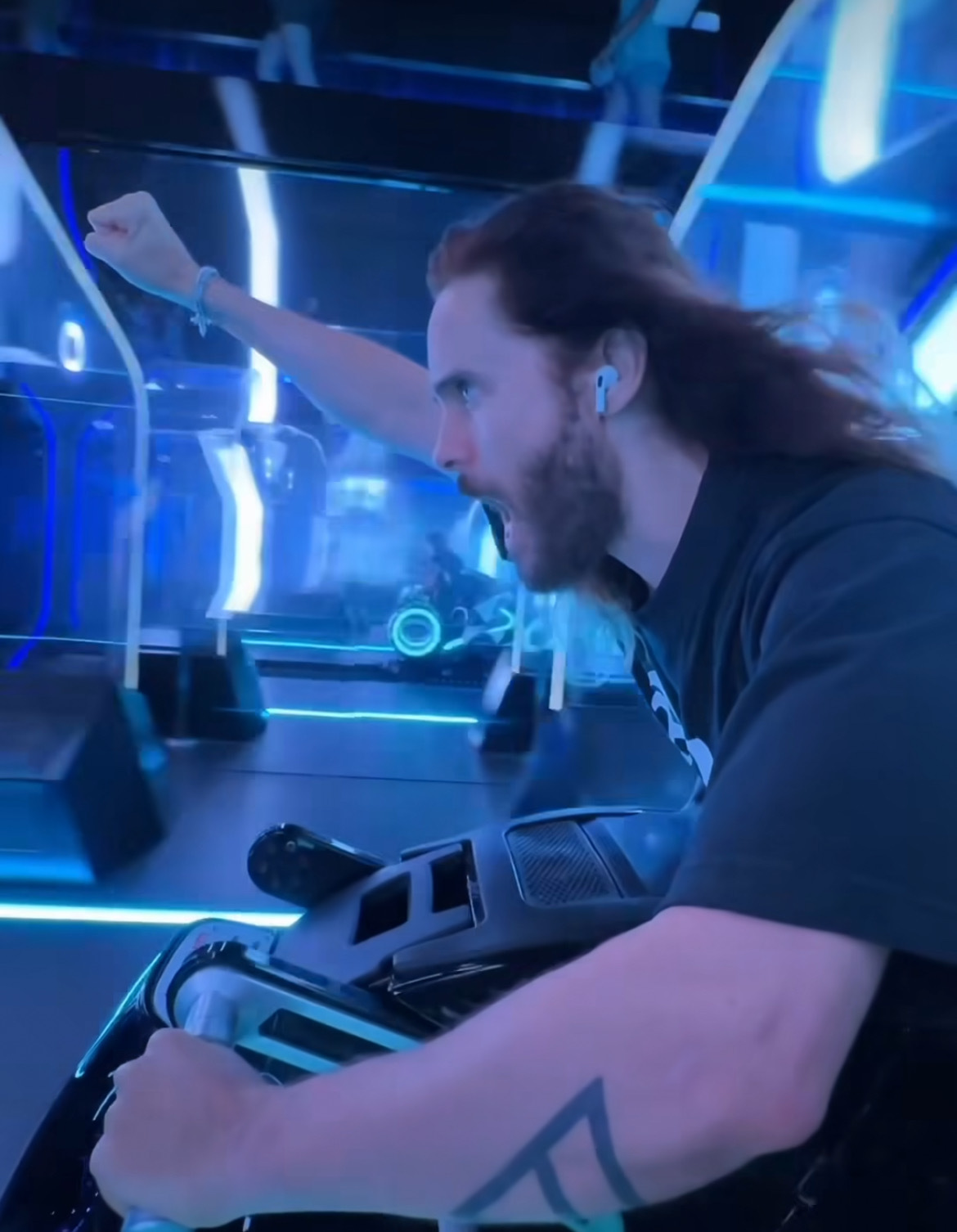 A person with long hair, wearing a black shirt and earbuds, poses energetically like Jared Leto while riding a futuristic, neon-lit motorcycle simulator indoors.