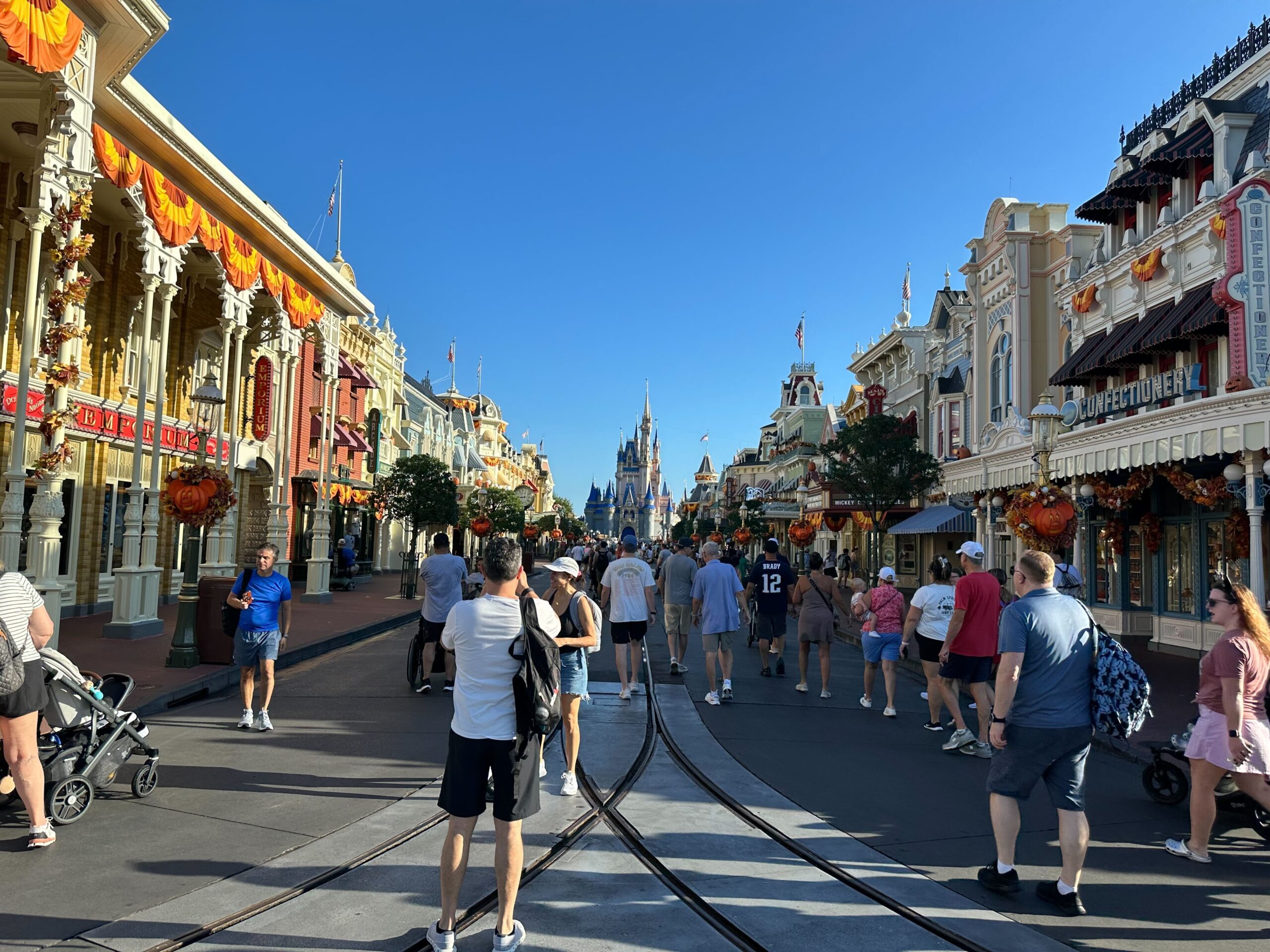 A bustling crowd walks down a decorated Main Street with shops on both sides and a castle visible in the distance under a clear blue sky.