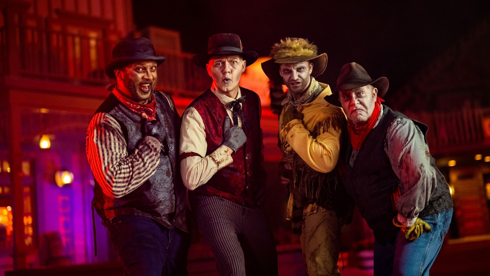 Four people dressed in cowboy attire pose in a Western-themed setting, with buildings illuminated in the background, evoking the charm of the Old West reminiscent of the Cadaver Dans.