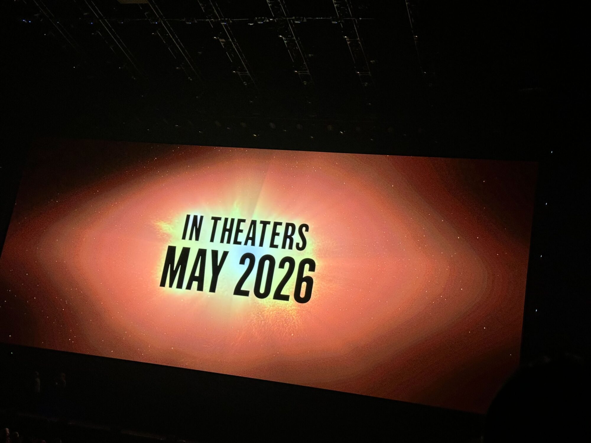 A large screen displays the text "In Theaters May 2026" against a backdrop of an orange and yellow abstract design, reminiscent of a vibrant scene from The Mandalorian.
