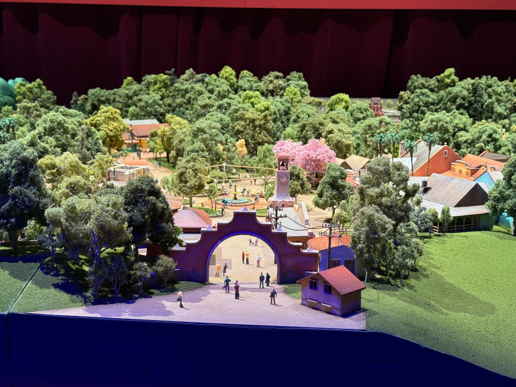 A detailed diorama features a miniature park with trees, a clock tower, buildings, and figurines. An archway serves as an entrance to what appears to be the idyllic village of Pueblo Esperanza, and the scene is surrounded by dense foliage.