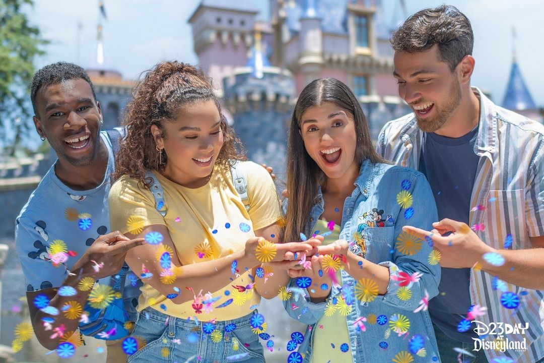Four people smiling and interacting with colorful confetti outside a theme park castle. The image is labeled with a "Disneyland" logo in the lower right corner.