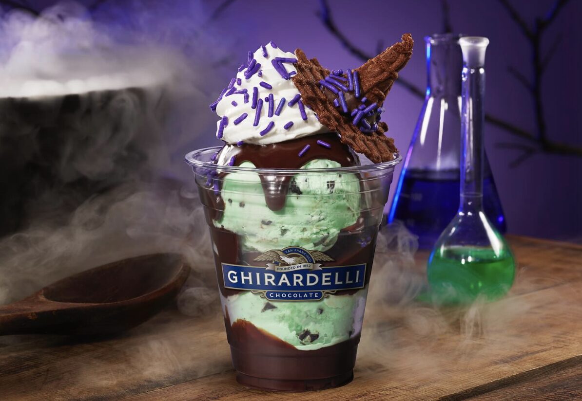 A dessert cup labeled "Ghirardelli" filled with mint chocolate chip ice cream, chocolate sauce, whipped cream, purple sprinkles, and a waffle cone piece resembling a witch's hat. This 'Hocus Pocus' Sundae is perfect for any fan visiting Disney California Adventure.