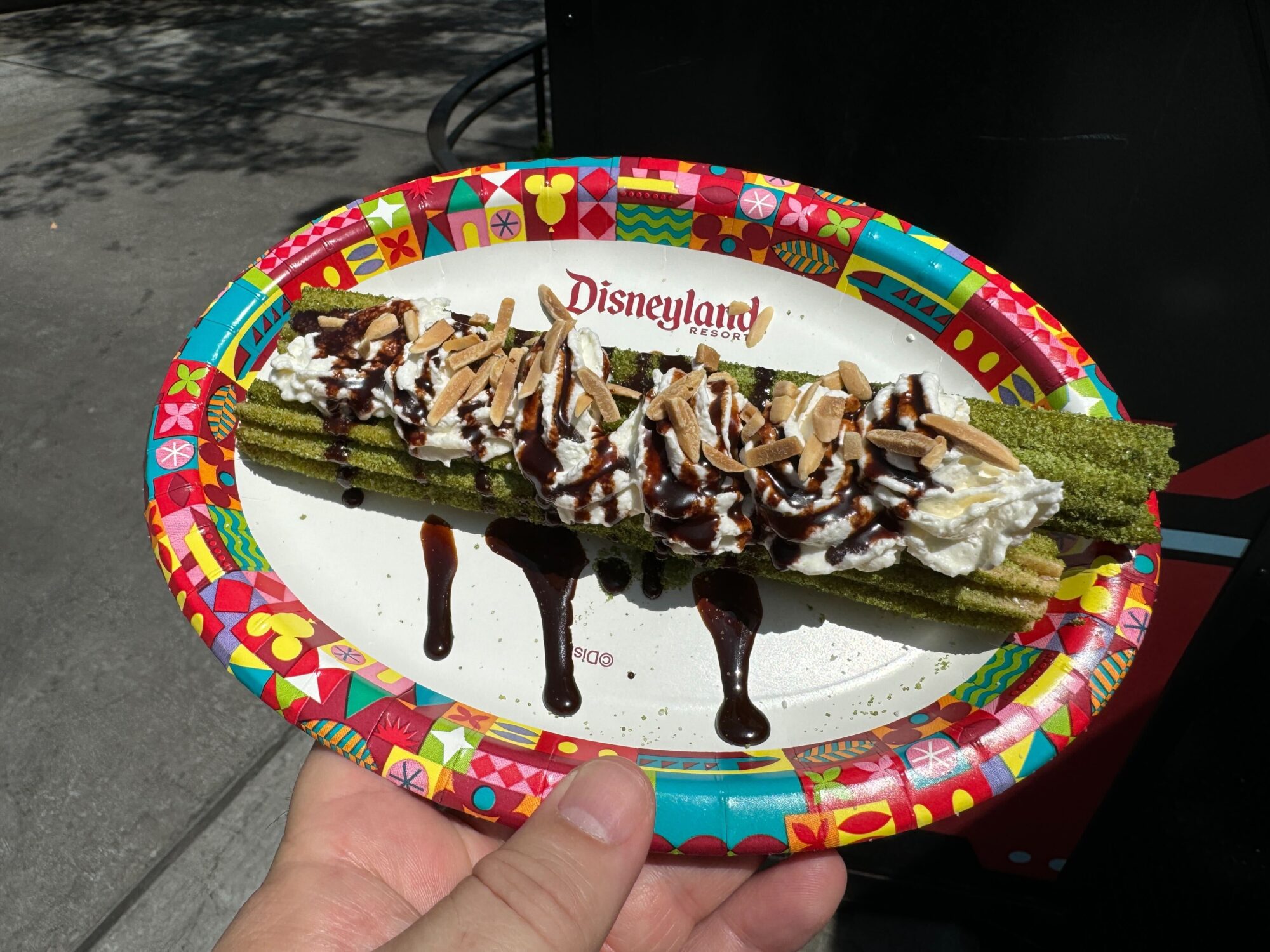 A hand holds a colorful plate with two green churros topped with whipped cream, chocolate drizzle, and almonds. The plate reads "Disneyland Resort.