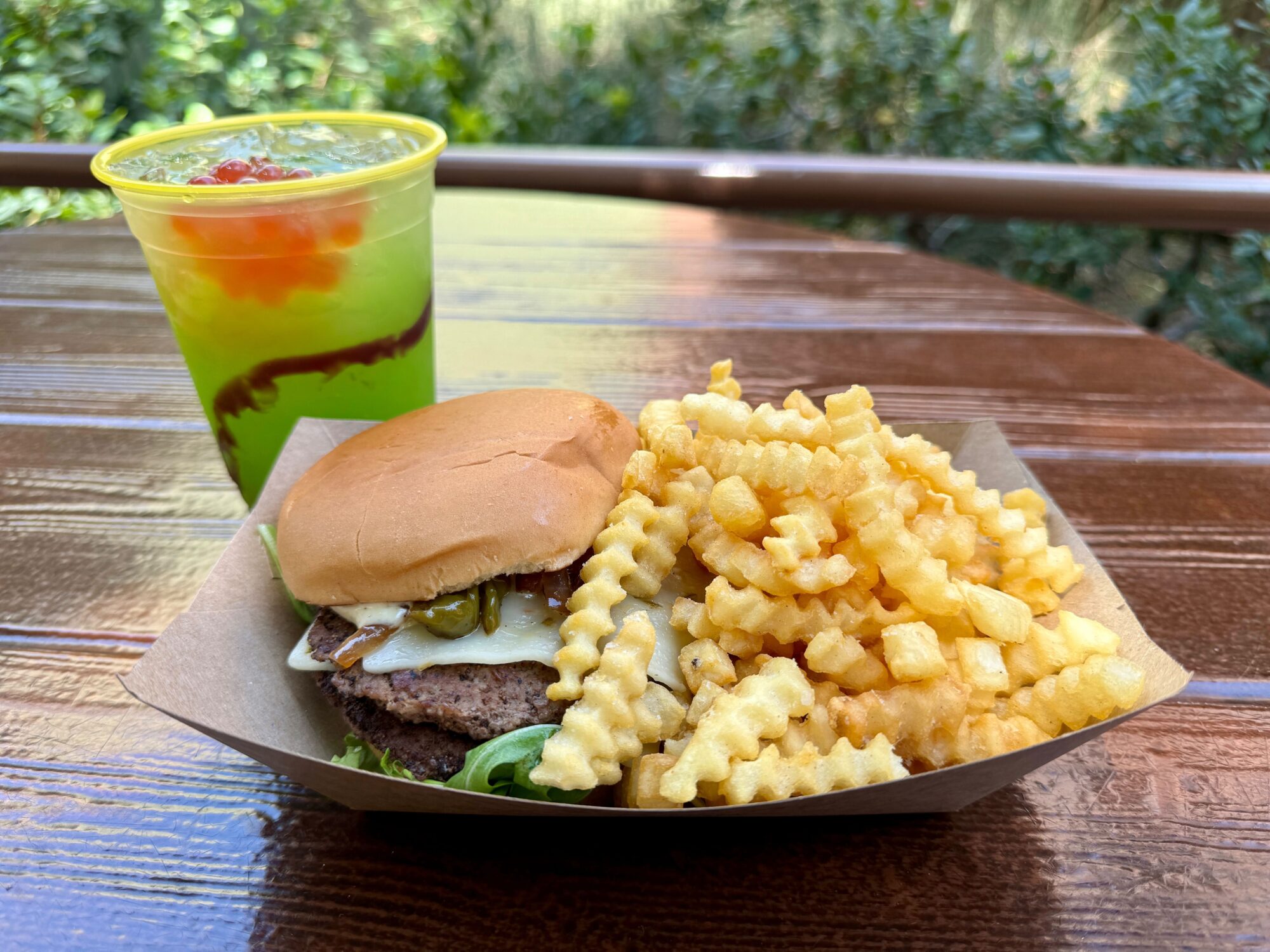 A cheeseburger with lettuce and jalapeños, crinkle-cut fries, and a green drink with red toppings are served on a wooden table, ready to refuel even the hungriest smokejumpers after a day of aerial firefighting.