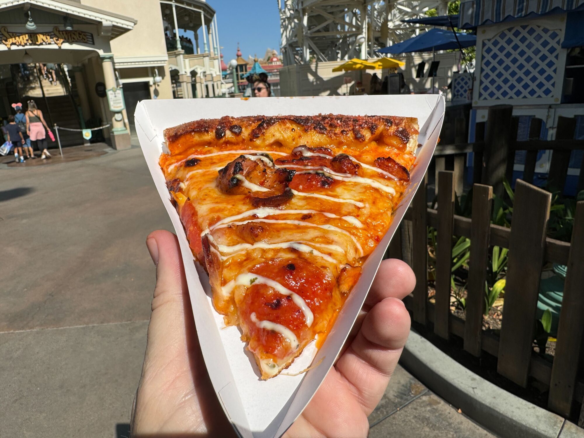 A hand holding a slice of spicy meats pizza with pepperoni and drizzled with white sauce in a triangular paper dish at an outdoor setting with people and amusement park buildings in the background.