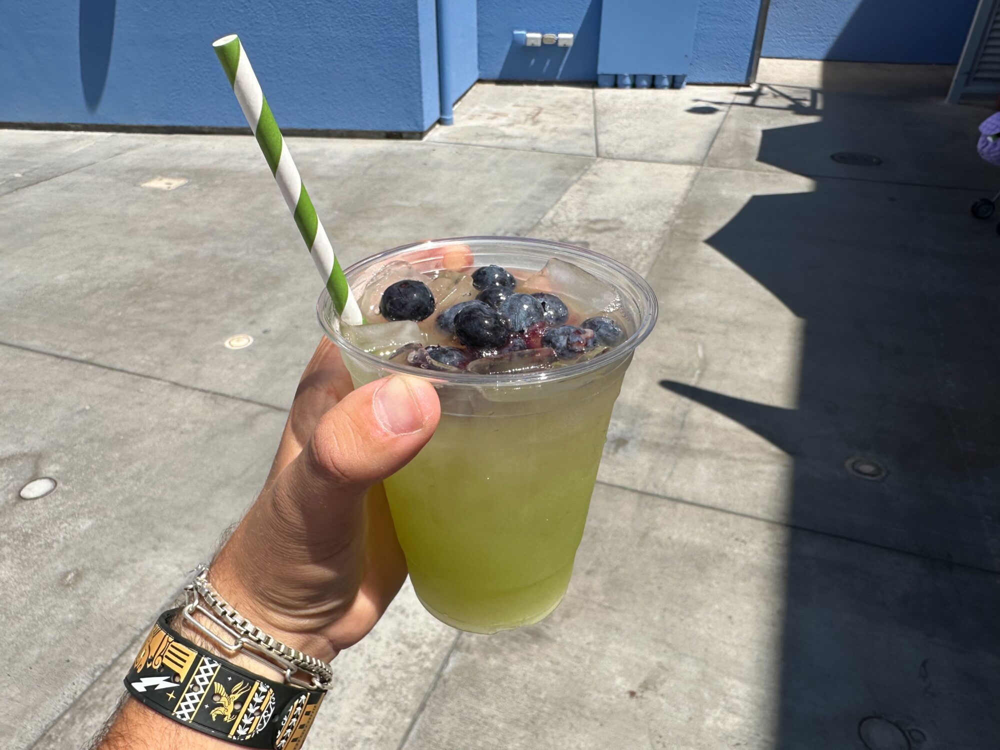 Hand holding a plastic cup filled with green-yellow liquid, ice, and blueberries. Cup has a green and white striped paper straw. Background shows a concrete outdoor area with shadows. The refreshing drink is a Cucumber-Mint Lemonade from Disney California Adventure.