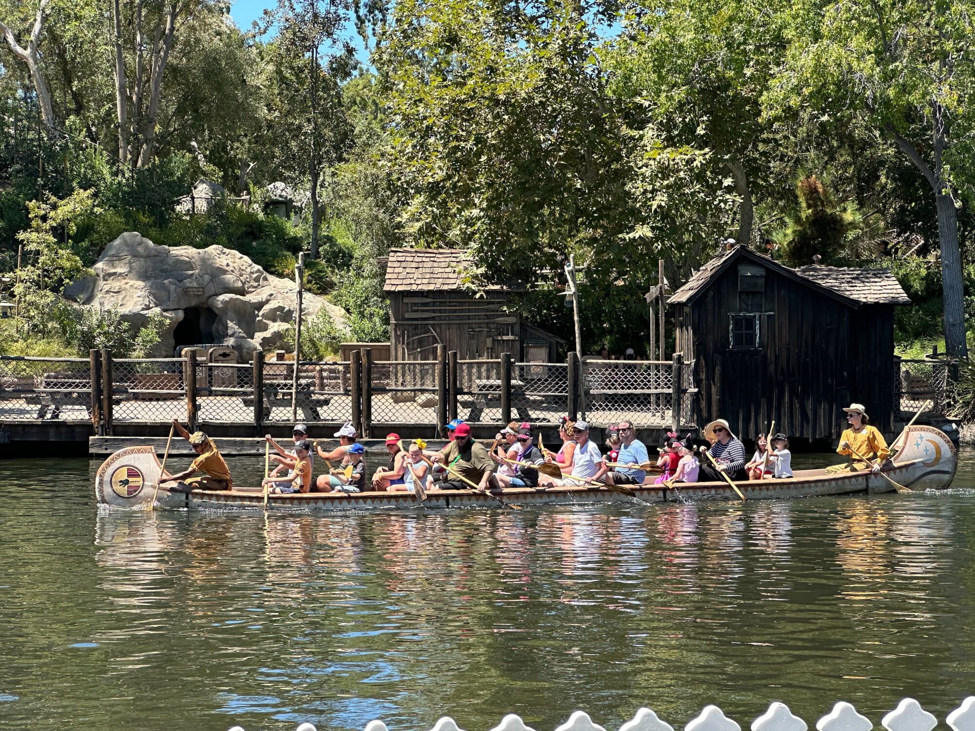A group of people in life vests paddle a canoe on a calm body of water. Trees and a rustic wooden cabin are visible in the background.