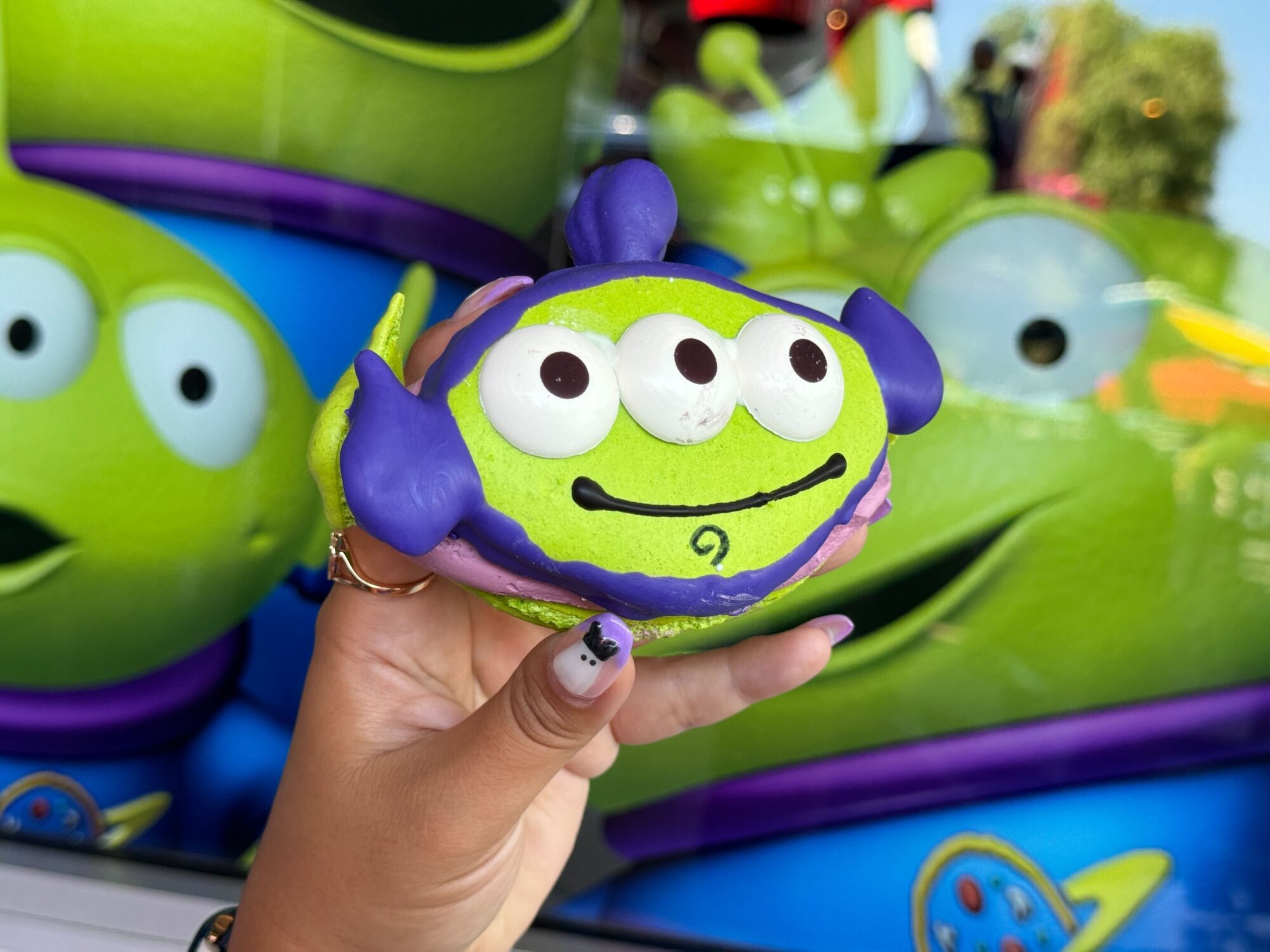 A hand holds a green alien-themed confectionery with three eyes and purple accents against a background featuring similar alien characters.