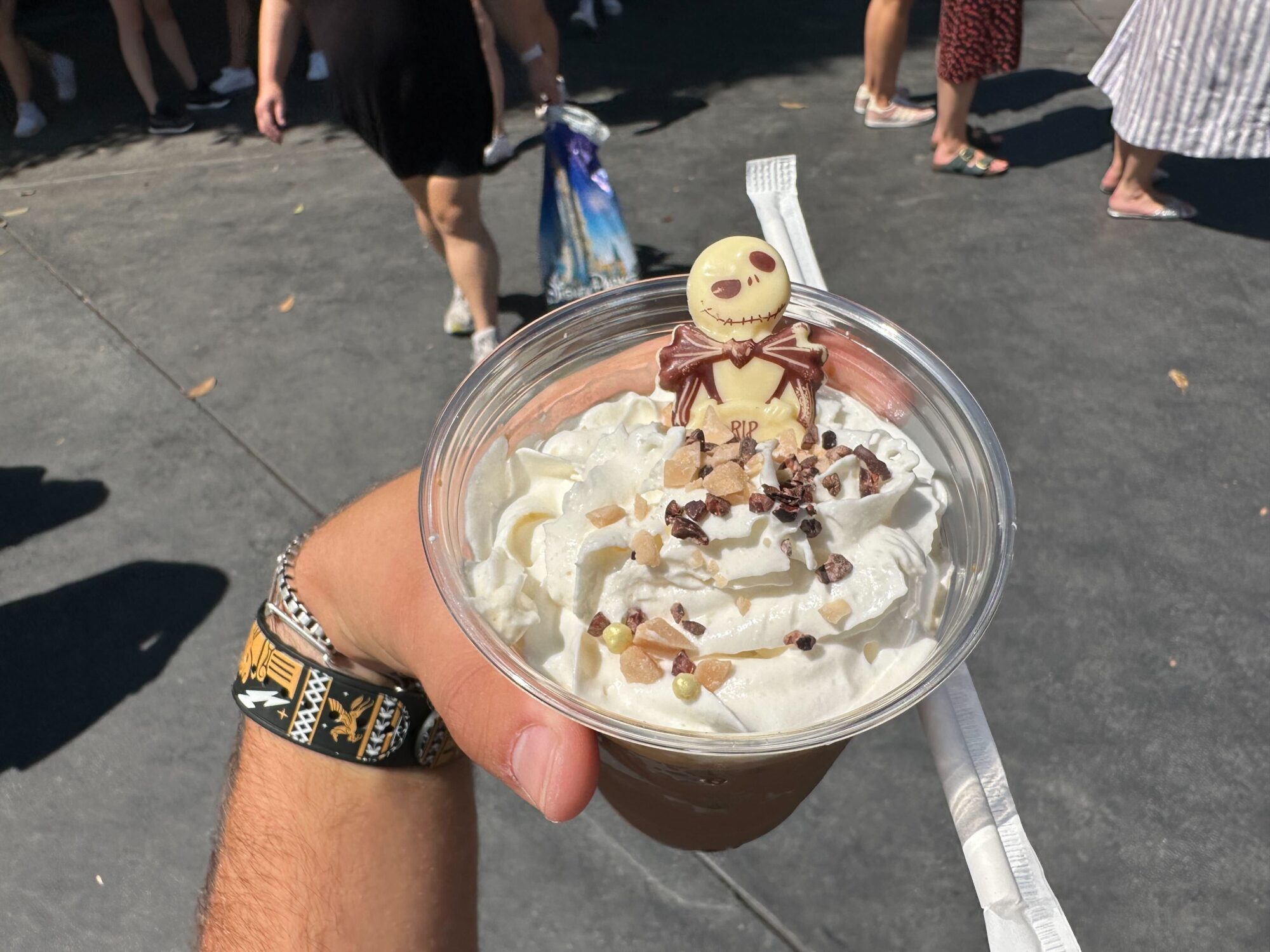 A hand holding a whipped cream-topped, pumpkin spiced dessert in a clear plastic cup with a decorative Jack Skellington chocolate piece. Other people walk by in the background.