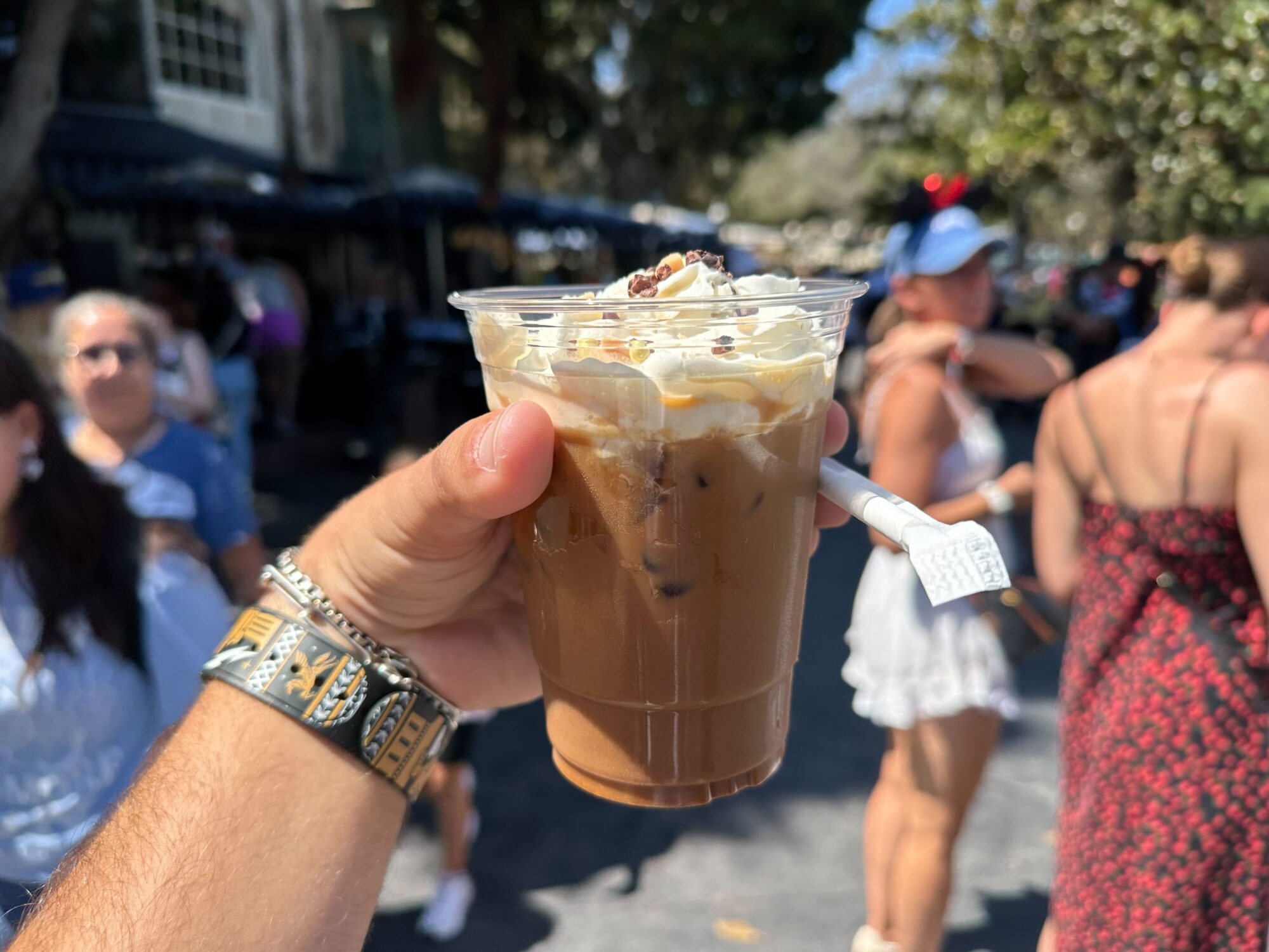 Person holding a plastic cup of pumpkin spiced iced coffee topped with whipped cream in an outdoor setting with people in the background.