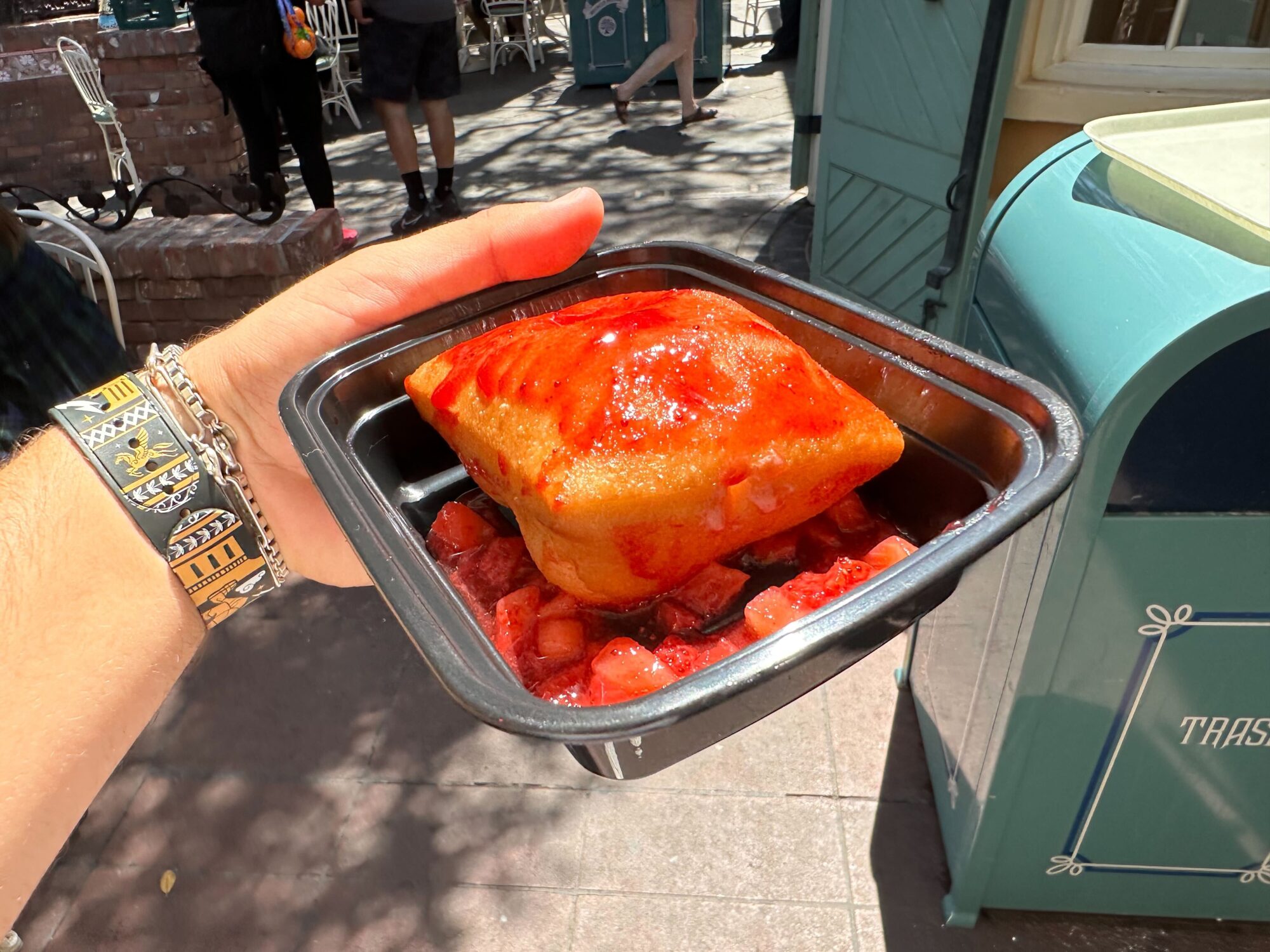 A hand holding a black plastic container with a square-shaped pastry topped with red sauce and diced strawberries, held up in outdoor sunlight. A blue trash bin is visible in the background.