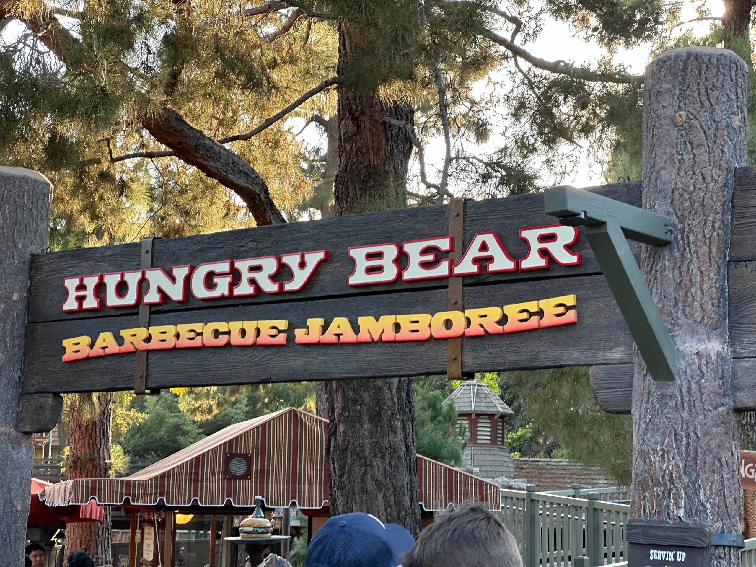 Sign above a restaurant entrance at Disneyland reads "Hungry Bear Barbecue Jamboree" with trees and a wooden structure visible in the background.