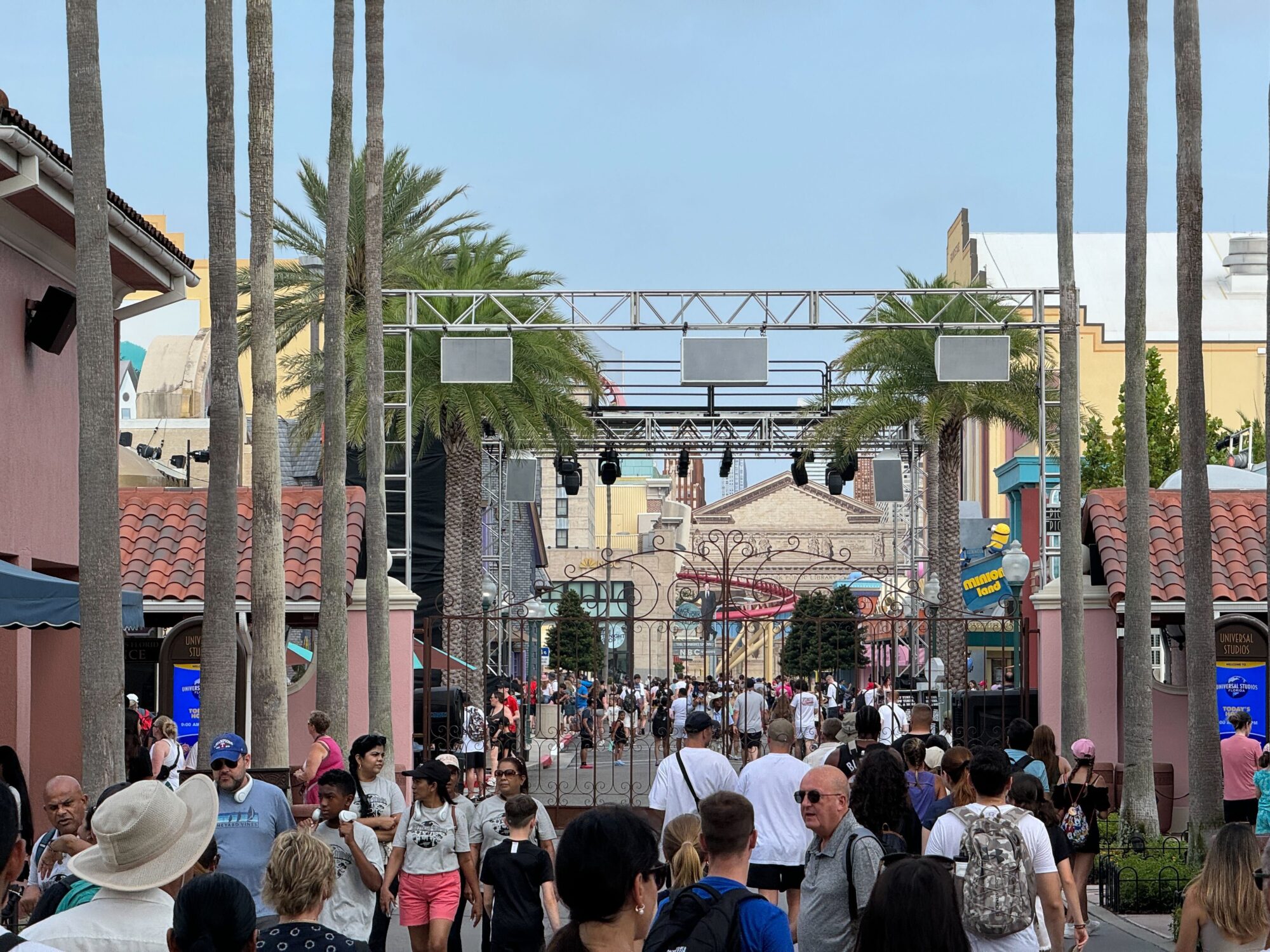 A busy theme park entrance with crowds of people, palm trees, and colorful buildings on either side. An overhead truss system with lights is visible in the foreground.