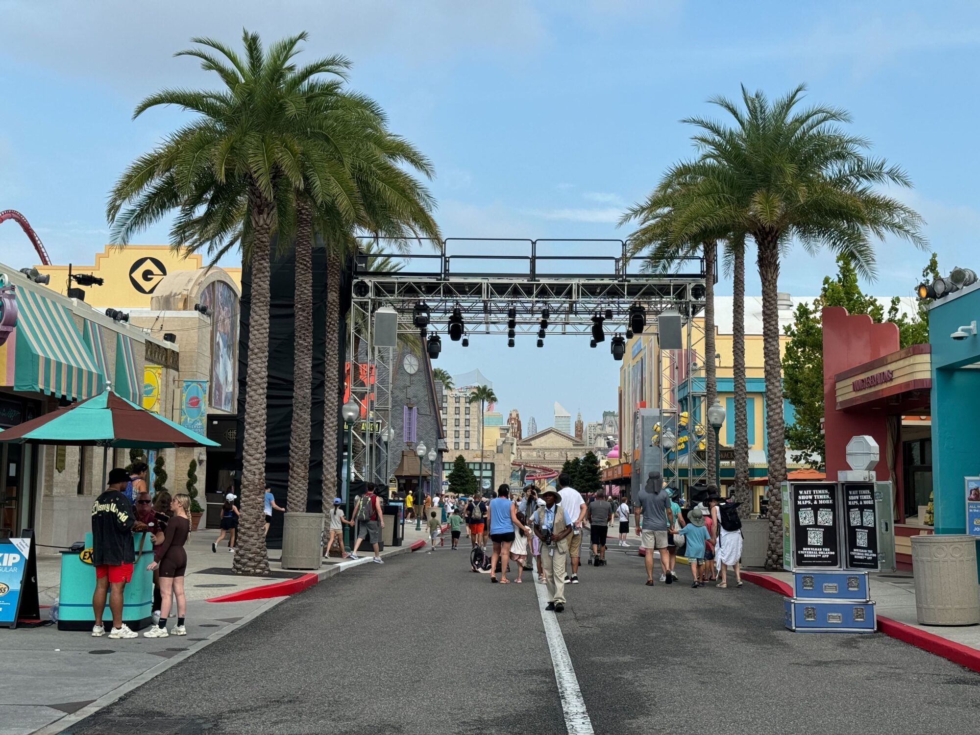 A city street with palm trees on either side, multiple colorful buildings, and people walking. Overhead is a metal truss structure with lights.