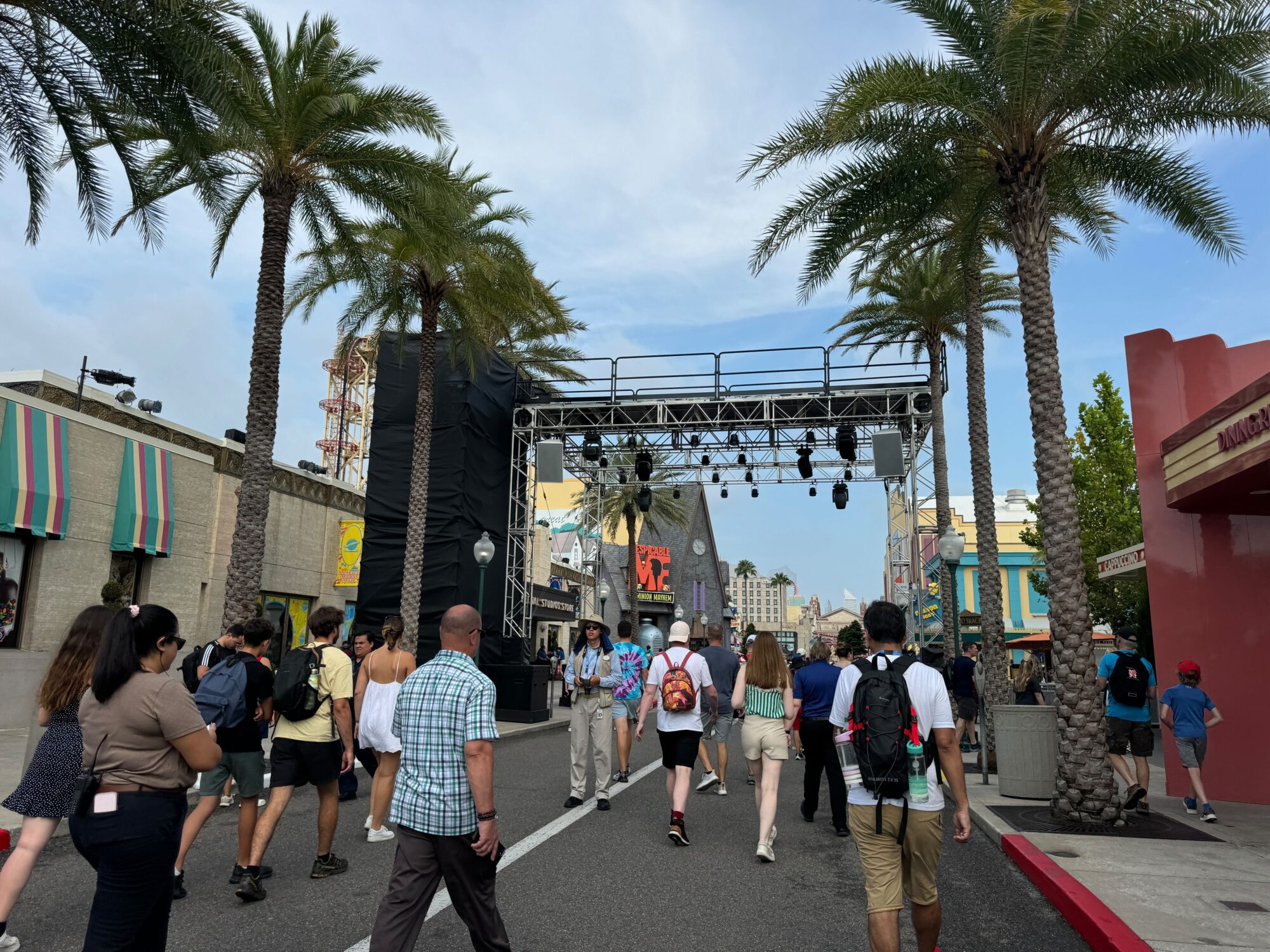 People walk along a palm tree-lined street with a stage and lighting rig set up in the background. The sky is partly cloudy.