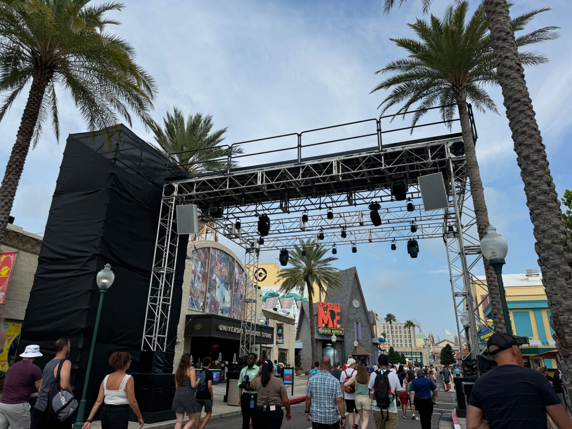 People walk along a palm tree-lined street under a large metal stage rigging with lights. There are various buildings in the background, including a theater with billboards.