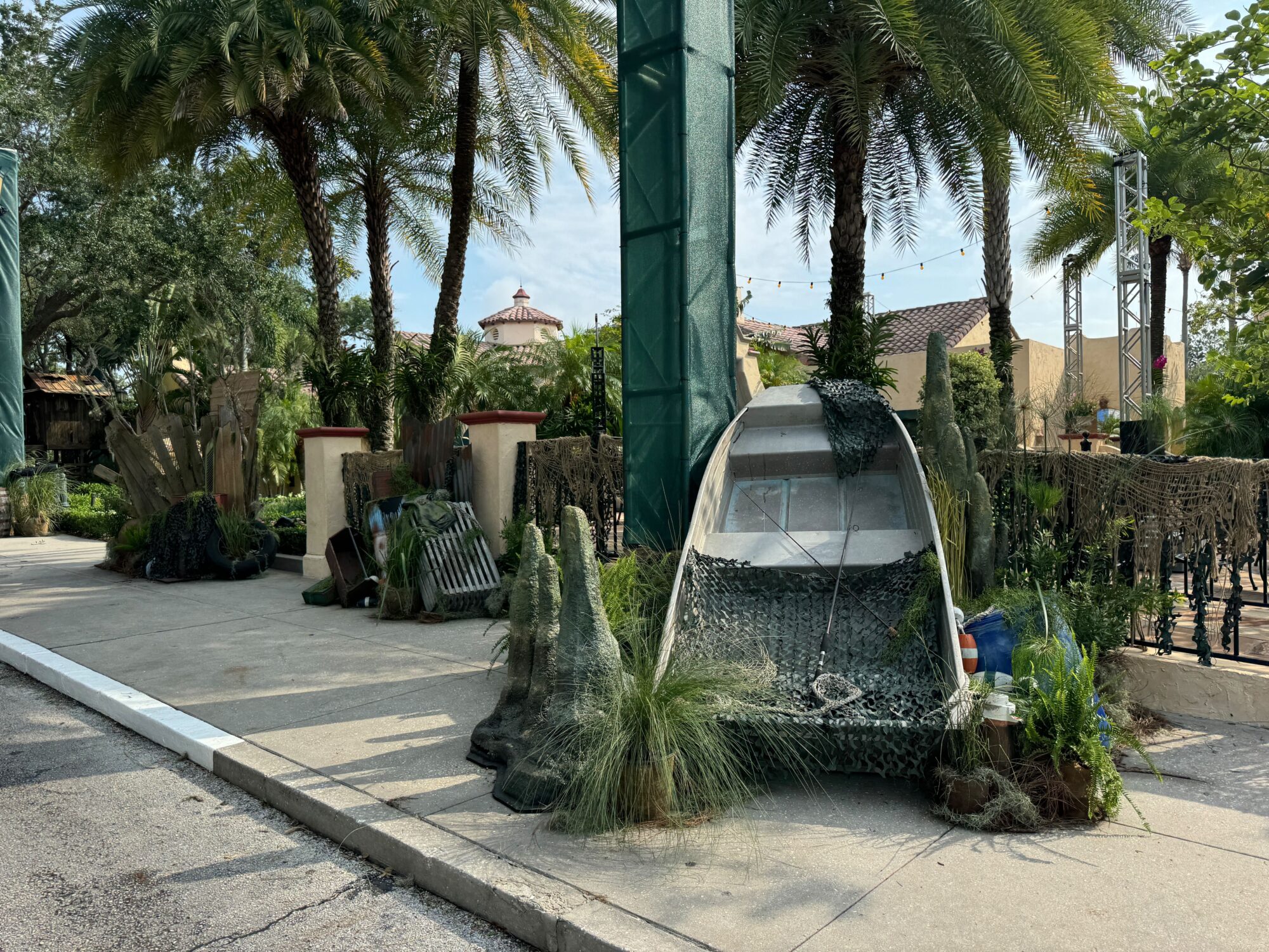 Decorative display with an old boat covered in netting, vegetation, and various props near palm trees and a pathway creates an immersive scare zone.