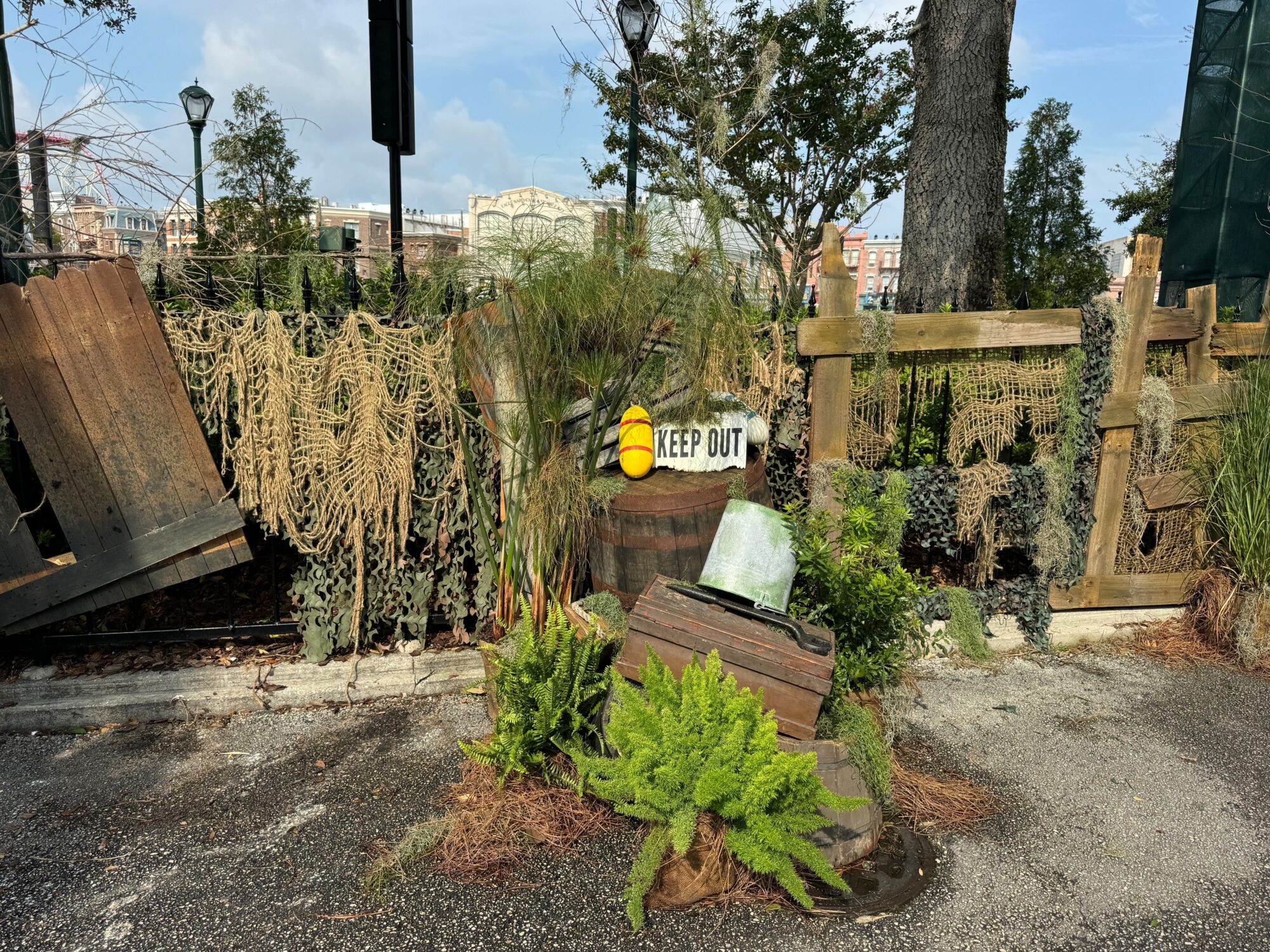 A weathered area with foliage, a "KEEP OUT" sign, old barrels, wooden crates, and netting forms a chilling scare zone set against a backdrop of trees and buildings.