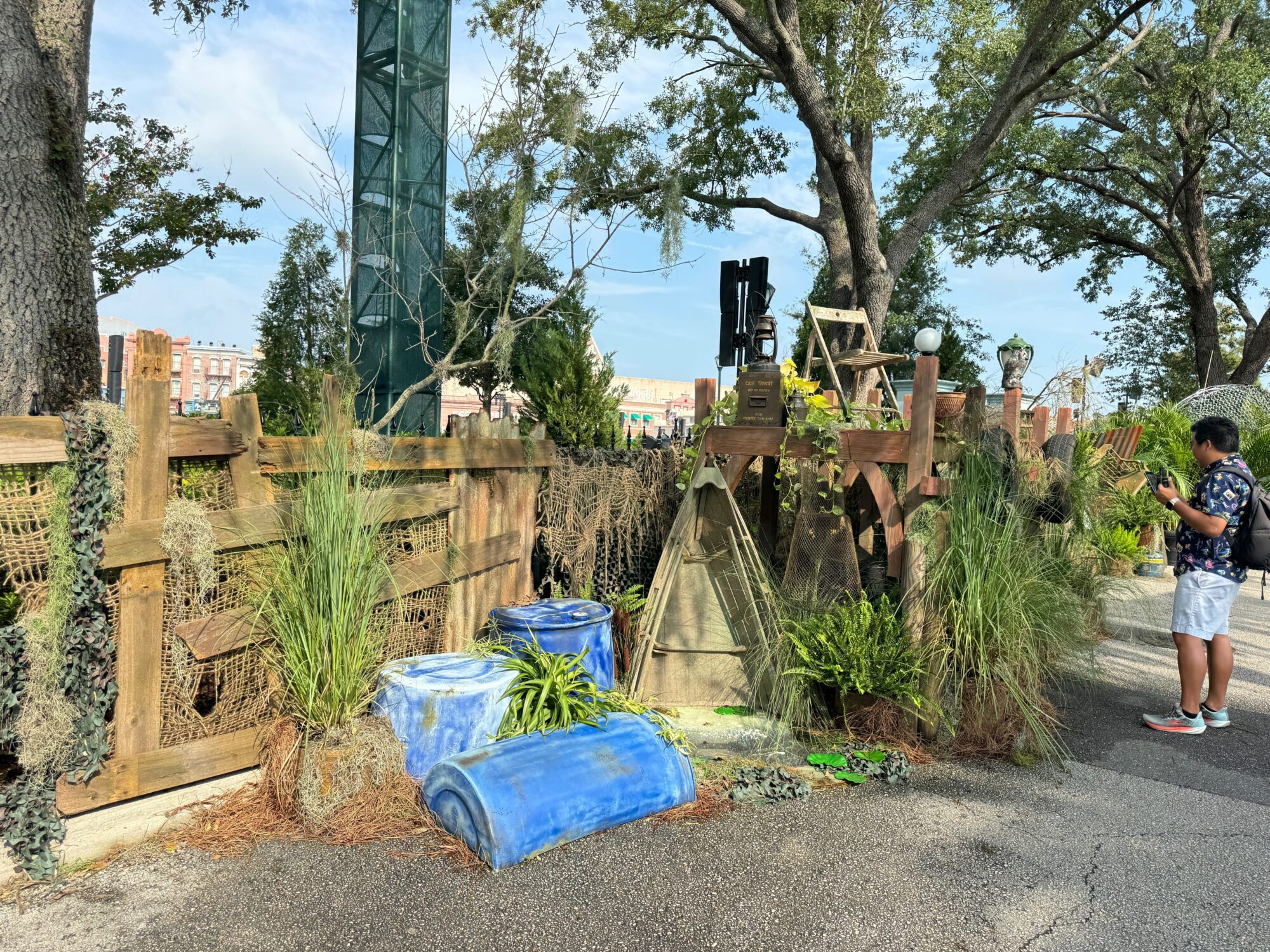 A man stands on the right side of the image near a rustic, decorative wooden fence adorned with foliage and two blue ceramic planters. Trees and a tall metal structure loom in the background, creating an eerie scare zone-like atmosphere.