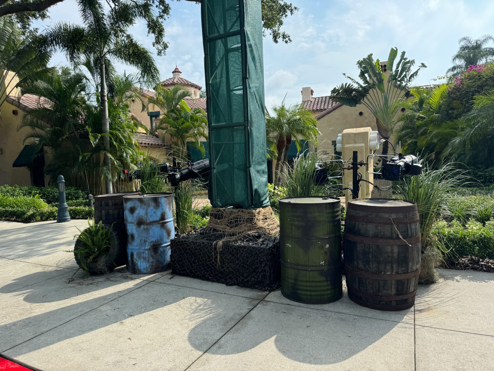 Barrels and drums are positioned around a roped-off, covered pole on a sunlit sidewalk, creating an eerie scare zone surrounded by tropical plants and trees.