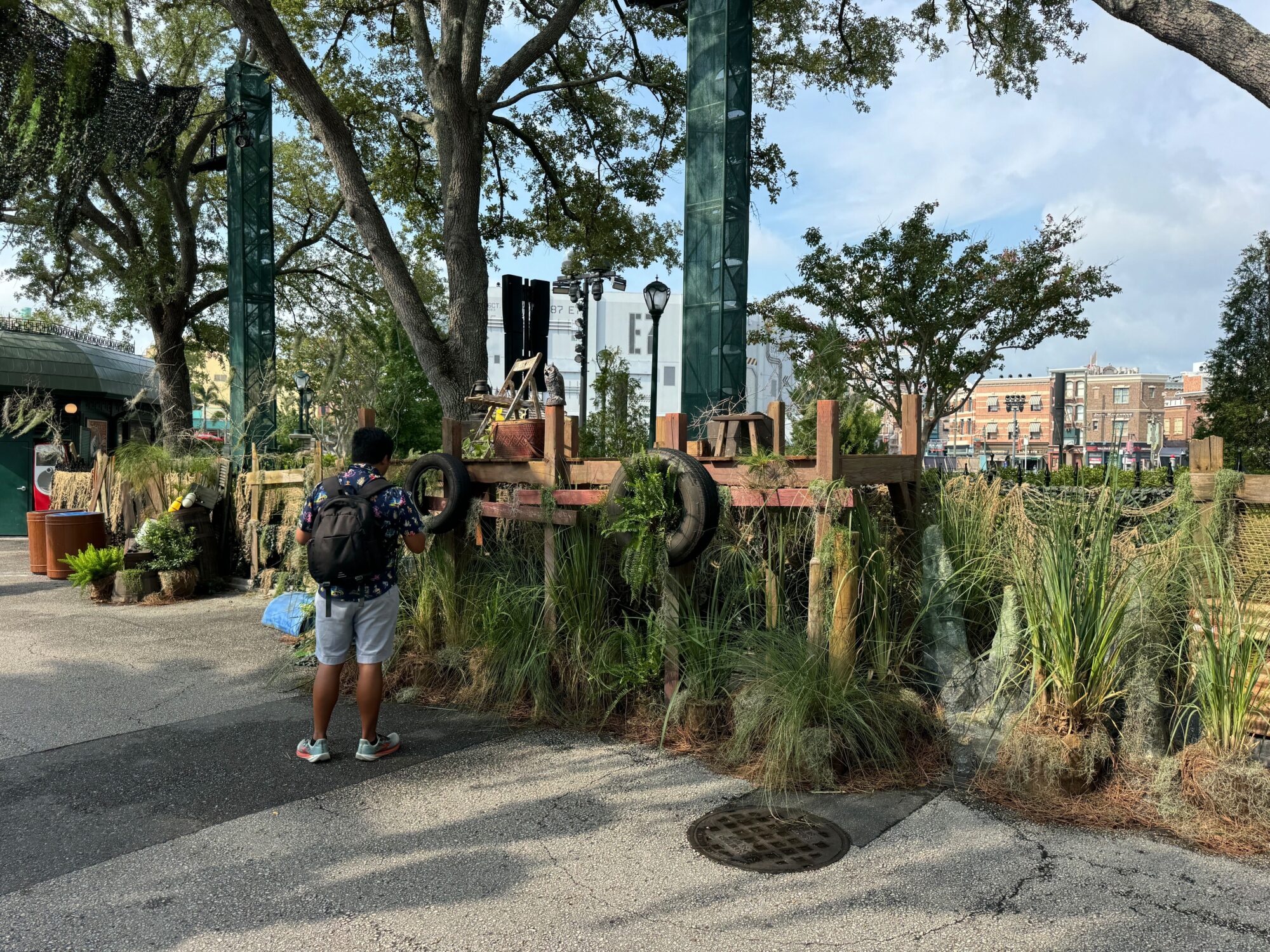 A person stands on a paved pathway near a wooden fence decorated with natural foliage and old tires, observing an outdoor installation in what appears to be a scare zone under a clear sky. Buildings are visible in the background.
