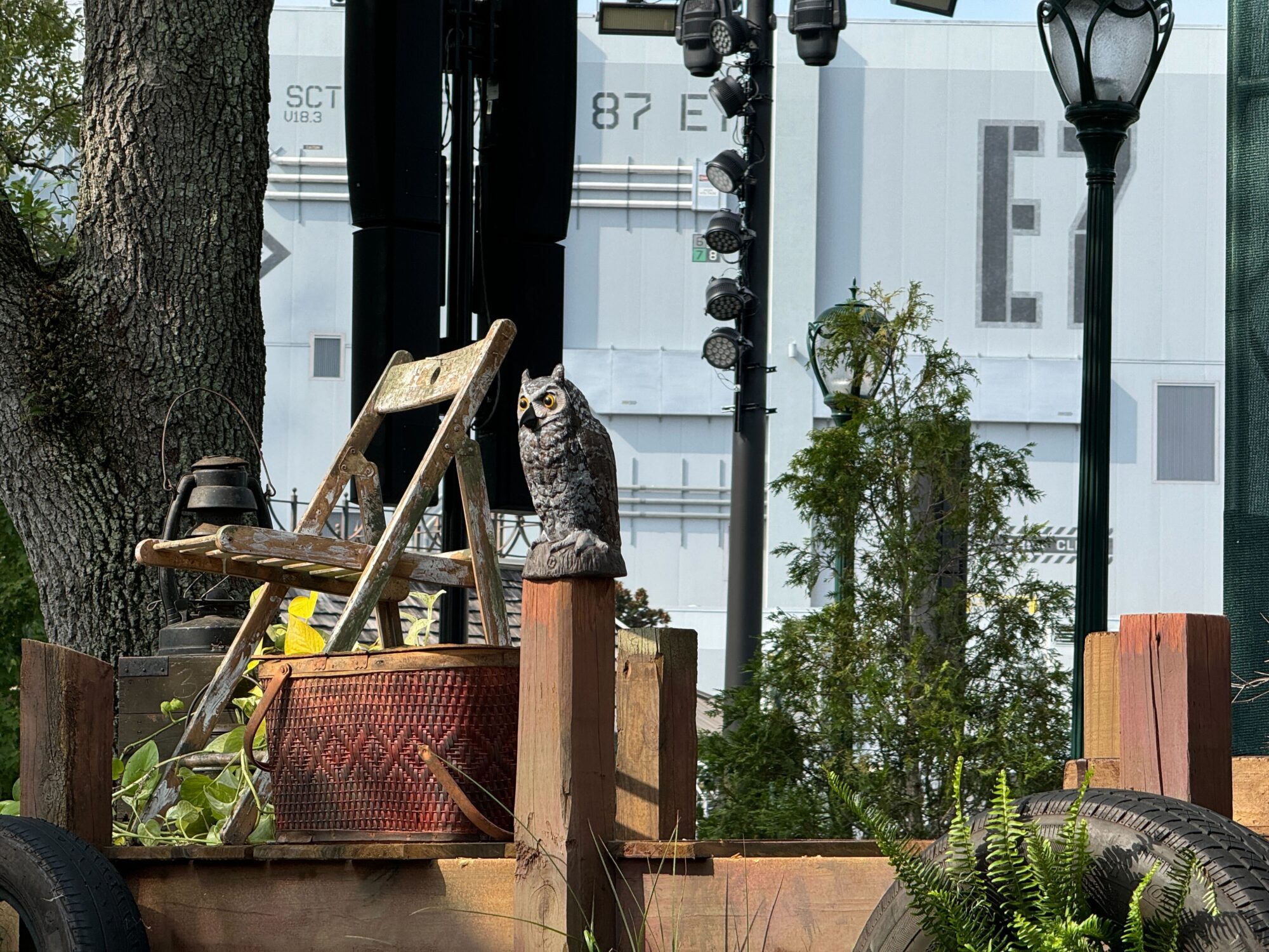 A scenic setup with a wooden chair, a wicker basket with foliage, and a lifelike owl statue, all placed on a wooden platform. In the background of this scare zone, there is industrial signage and greenery.