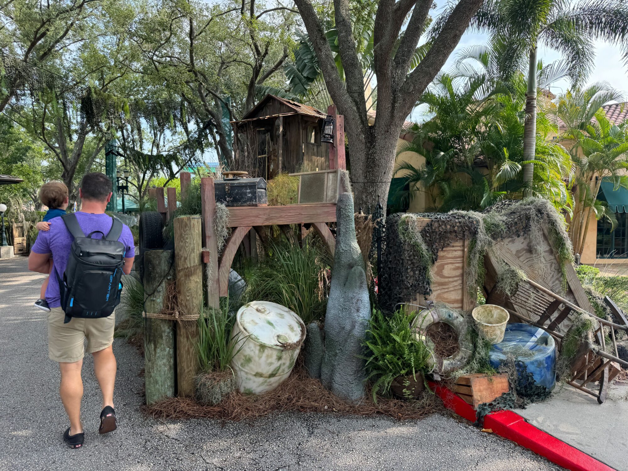 A man carries a child while walking past a cluttered outdoor setup with various barrels, wooden structures, and foliage. Trees and plants surround the scare zone, adding to its eerie atmosphere.