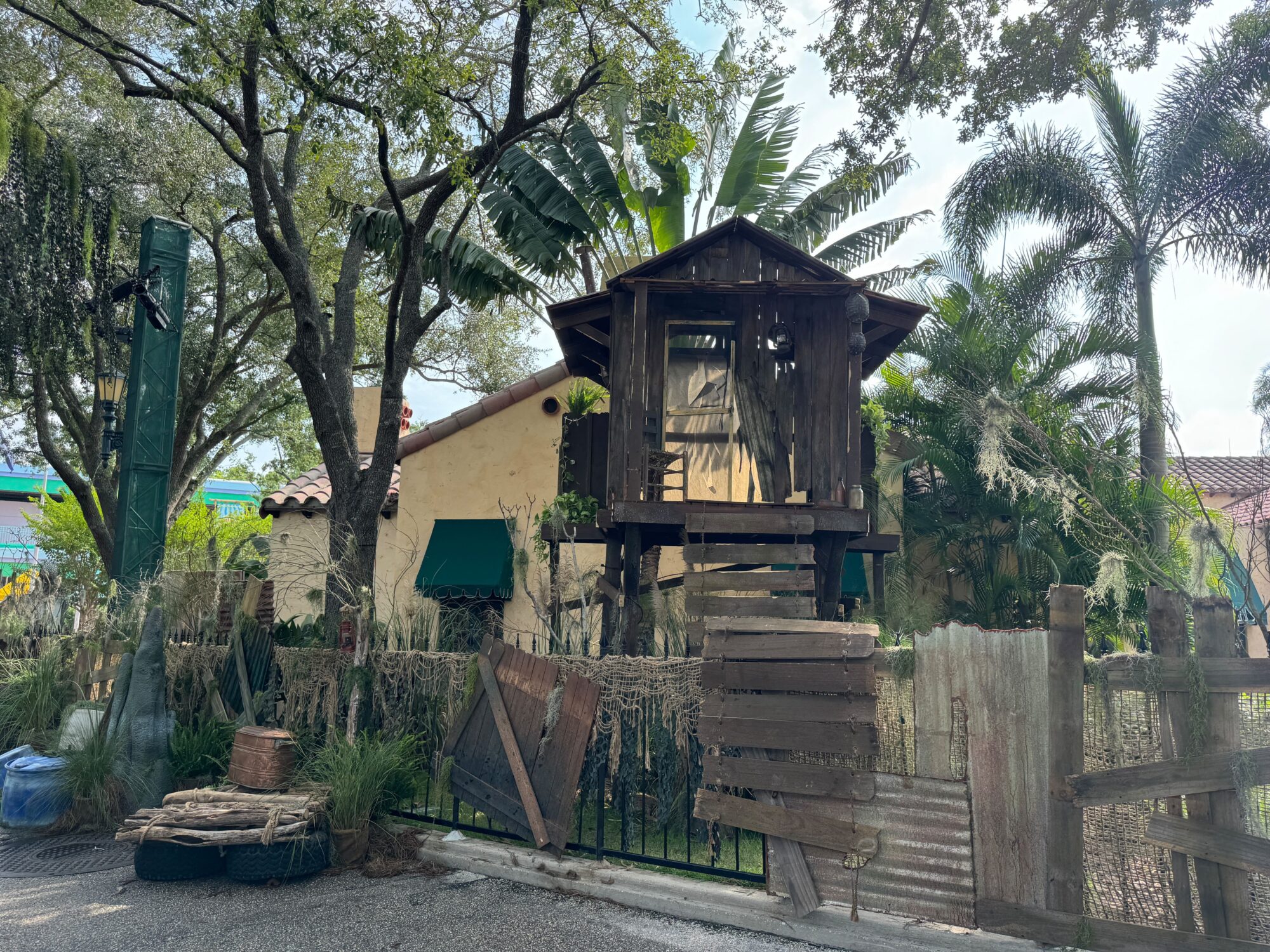 A treehouse with a wooden staircase is situated in a lush, tropical garden, surrounded by palm trees and various foliage. The ambiance resembles a tranquil retreat more than a scare zone. An adjacent house is partially visible in the background.