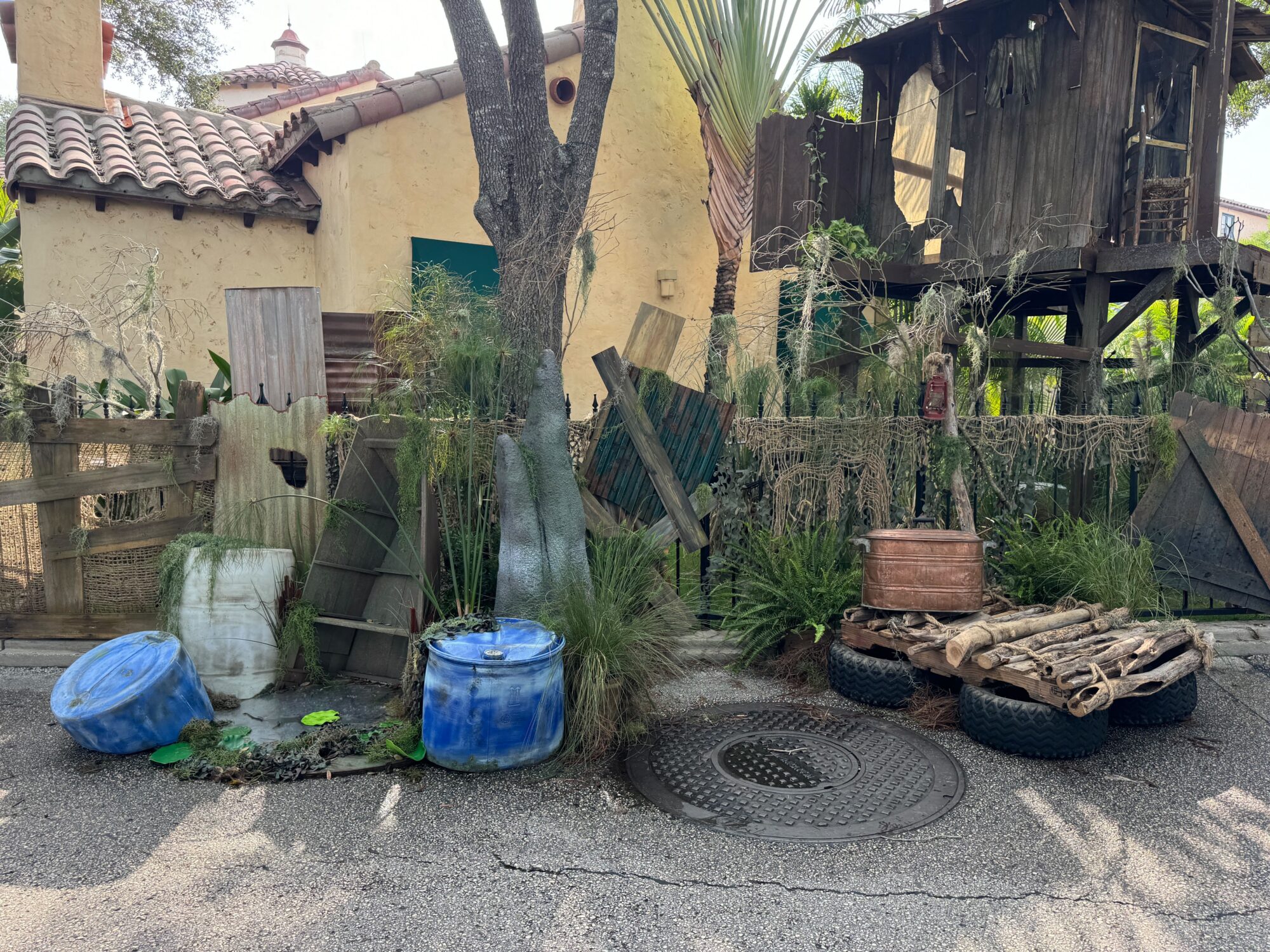A rustic scare zone scene features a treehouse, weathered barrels, metal containers, and plants. The ground has a manhole cover and wooden platform with tires. A stucco building forms the backdrop.
