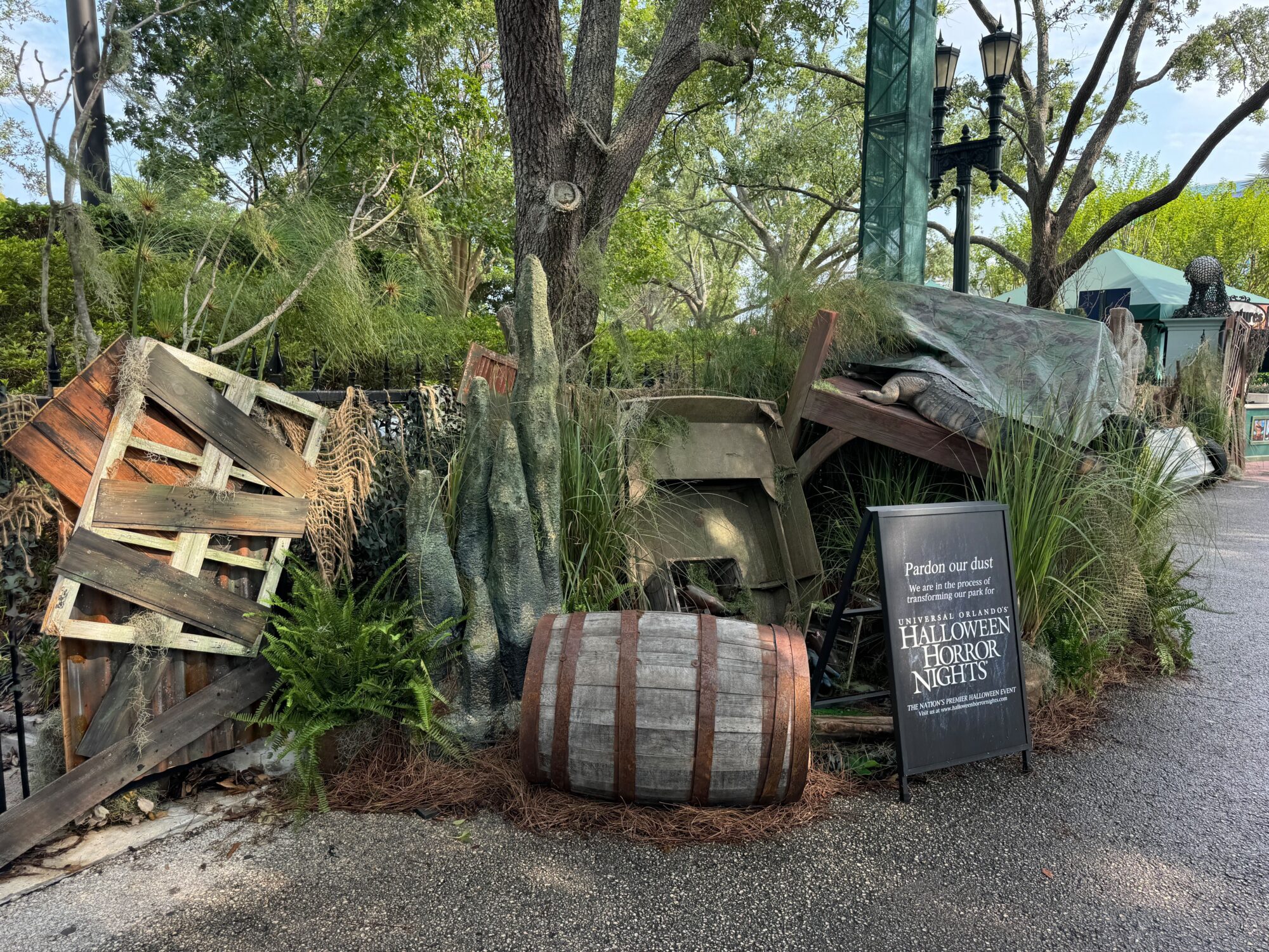 A pile of weathered crates, barrels, and wood beams is surrounded by various plants and greenery. A sign in the foreground reads "Pardon our dust Halloween Horror Nights scare zone.