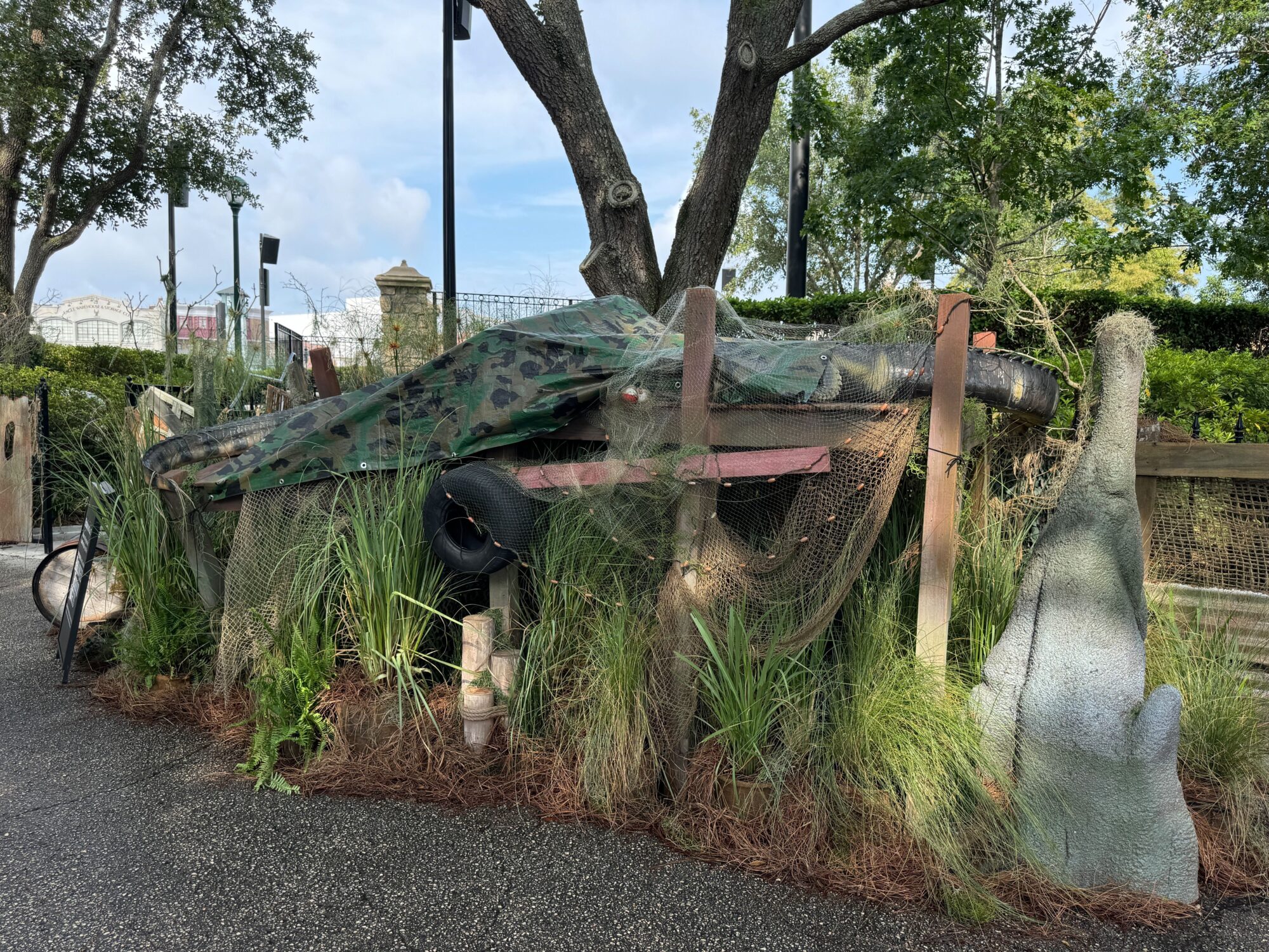 A small wooden structure covered with camouflage netting and surrounded by vegetation and various props sits in an outdoor scare zone. Trees and a section of a fence are visible in the background.