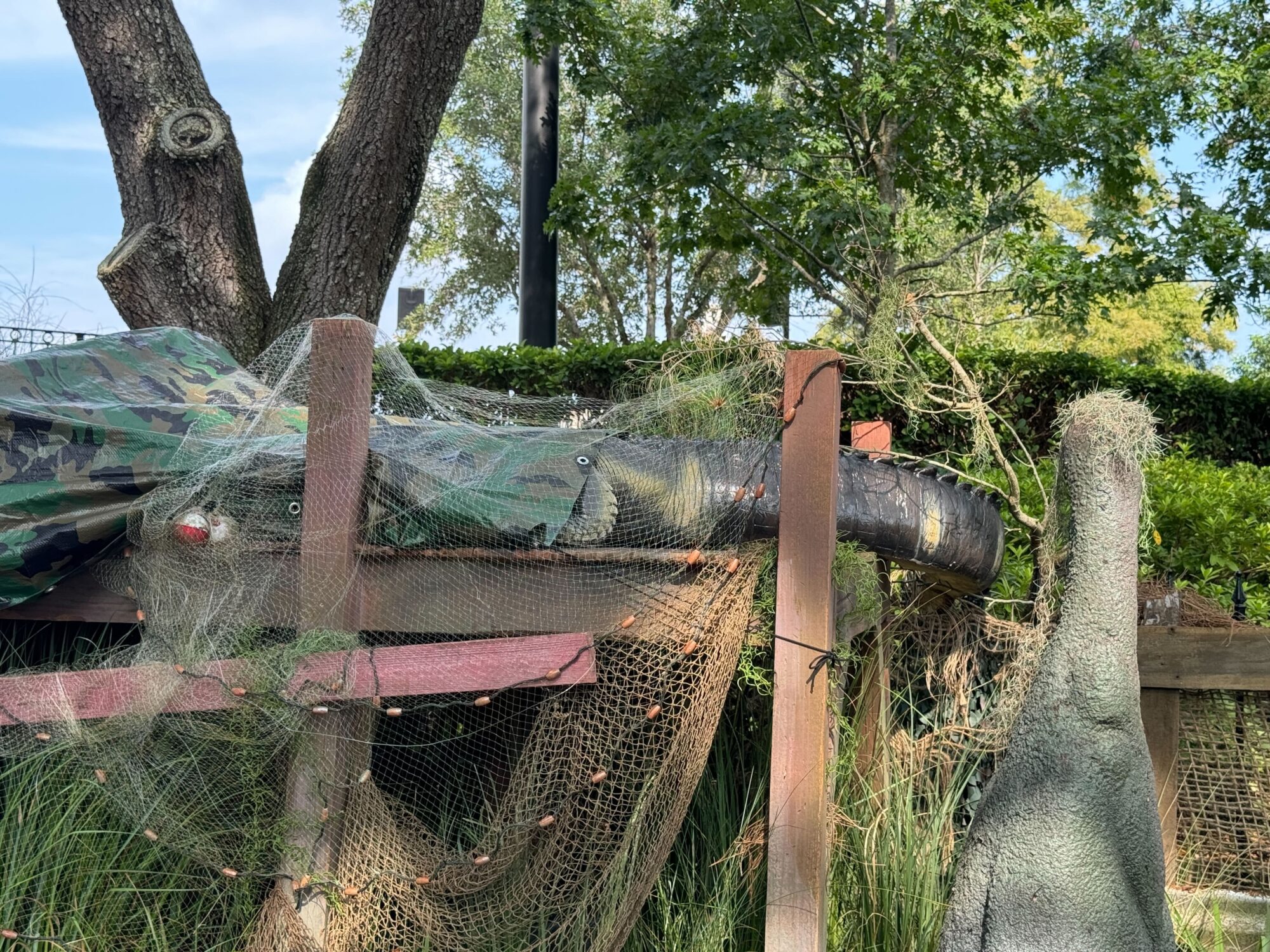 A camouflaged scare zone with netting and wood planks surrounding an animatronic dinosaur's tail and head among trees and greenery.